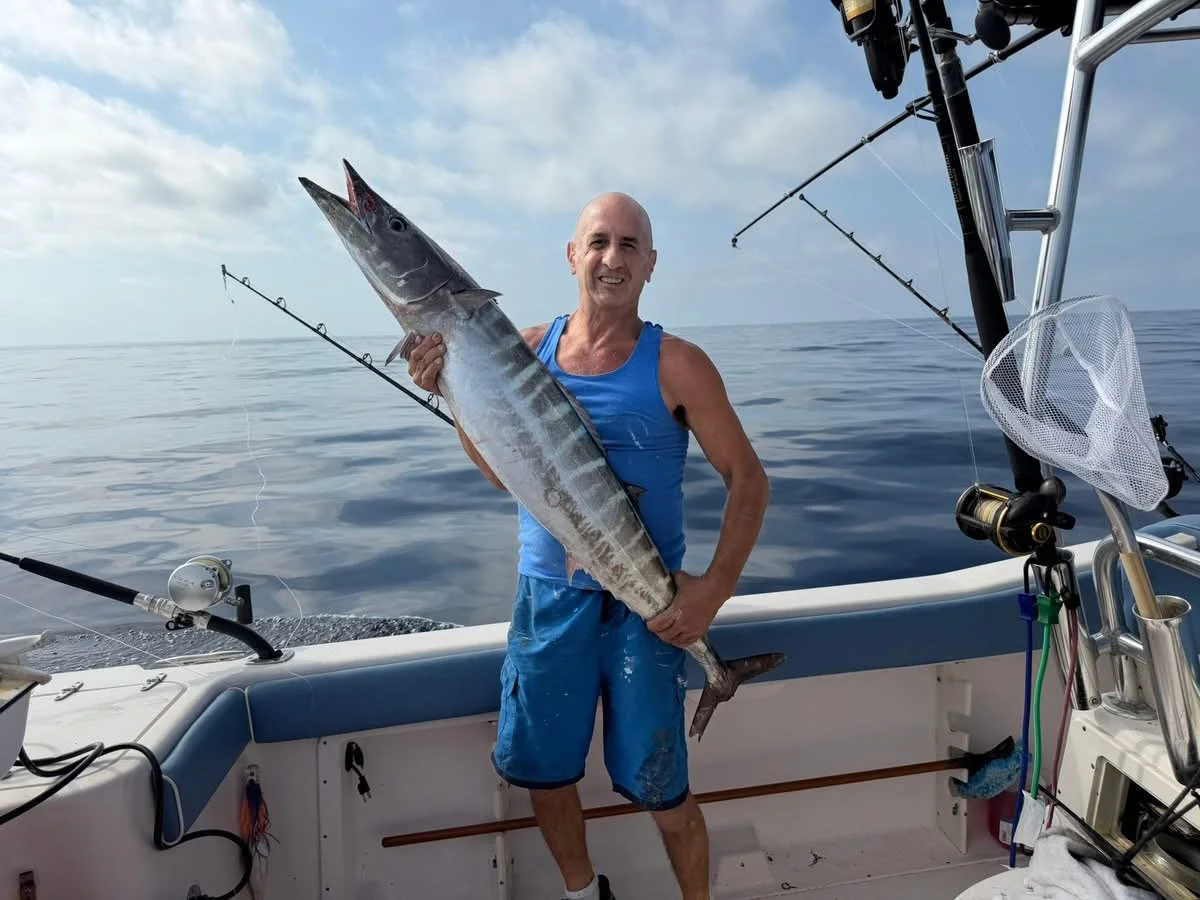 A man in a blue sleeveless shirt and shorts holding a large fish on a boat in open water, with fishing rods and equipment visible.