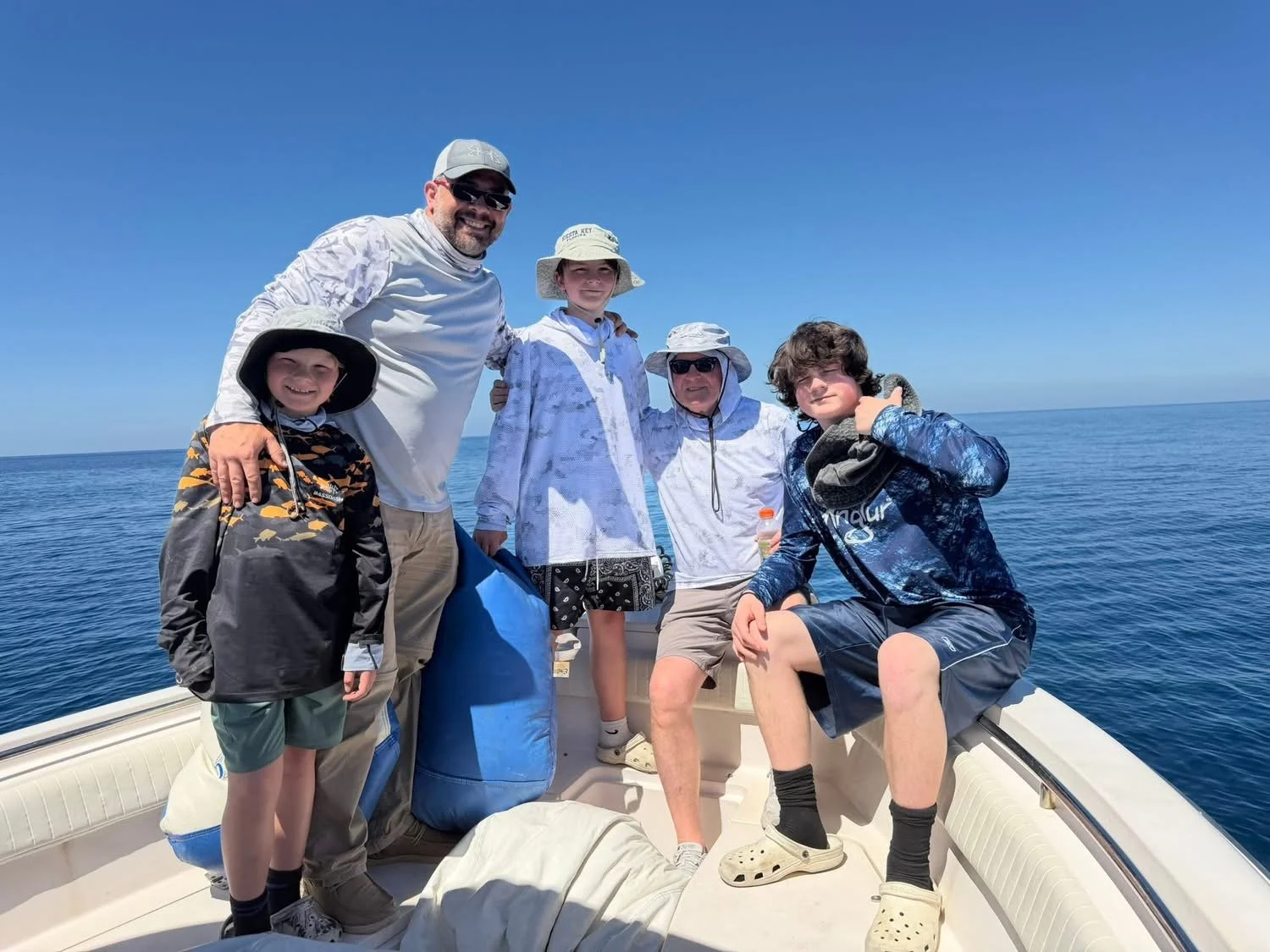 Group of five people on a boat in the ocean enjoying a sunny day, wearing hats and casual clothing.