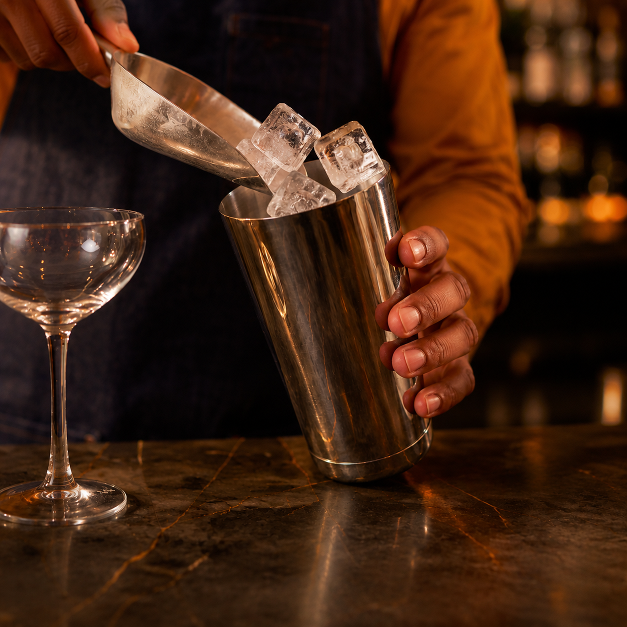 Person pouring ice into a metal cocktail shaker at a bar, with a cocktail glass nearby.