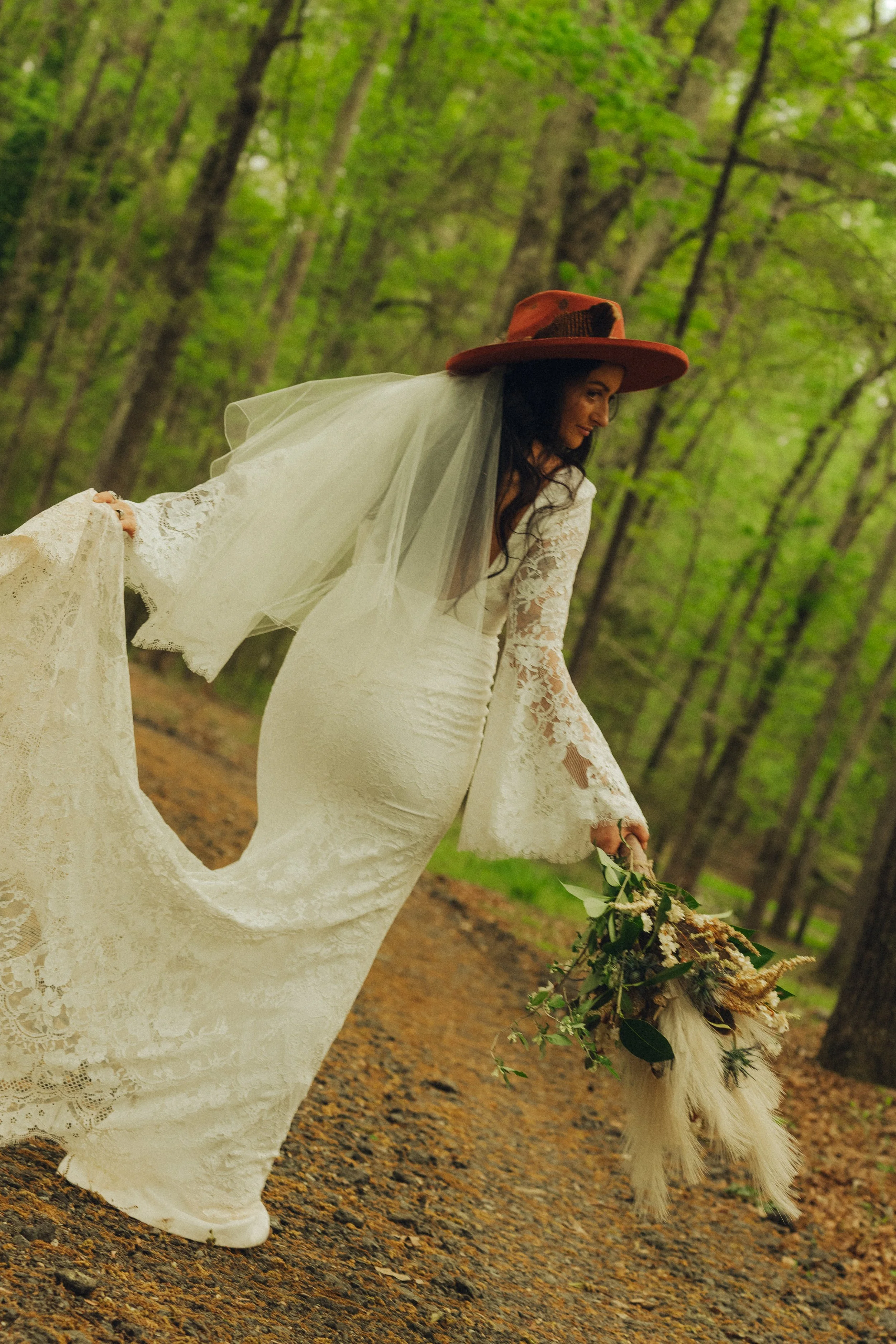 A woman wearing a white lace wedding dress and a large orange hat with a black pattern, walking on a forest path while holding a bouquet of flowers and a fluffy white dog.