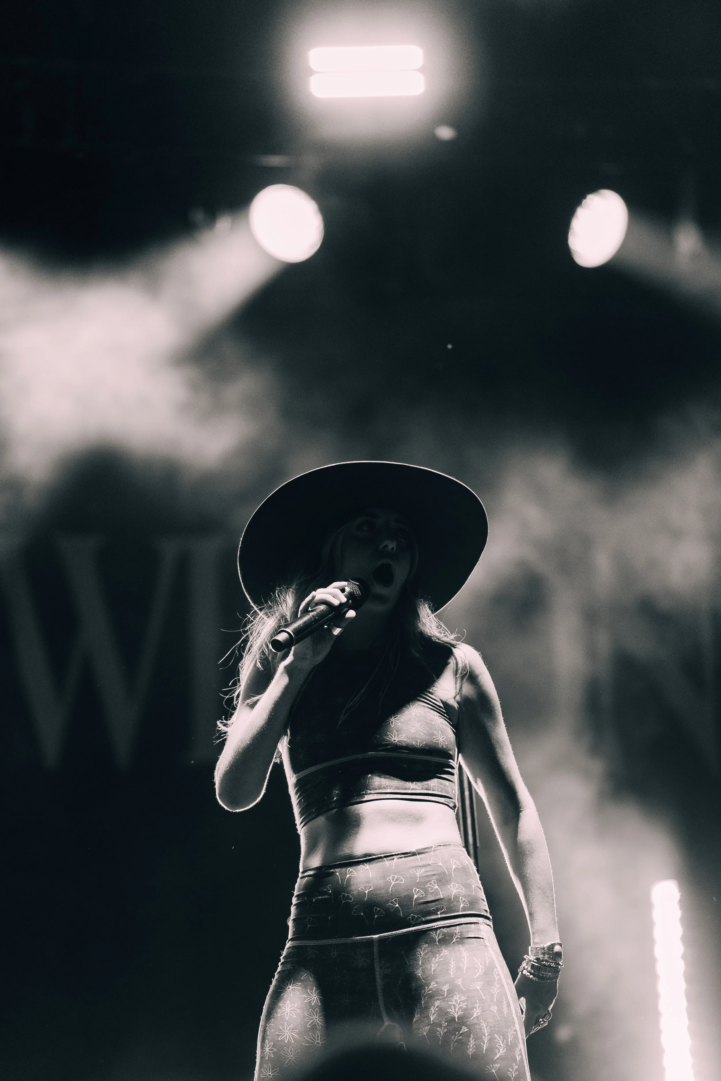 A woman singing into a microphone on stage, wearing a large hat and a patterned crop top, black and white photo with bright stage lights overhead.