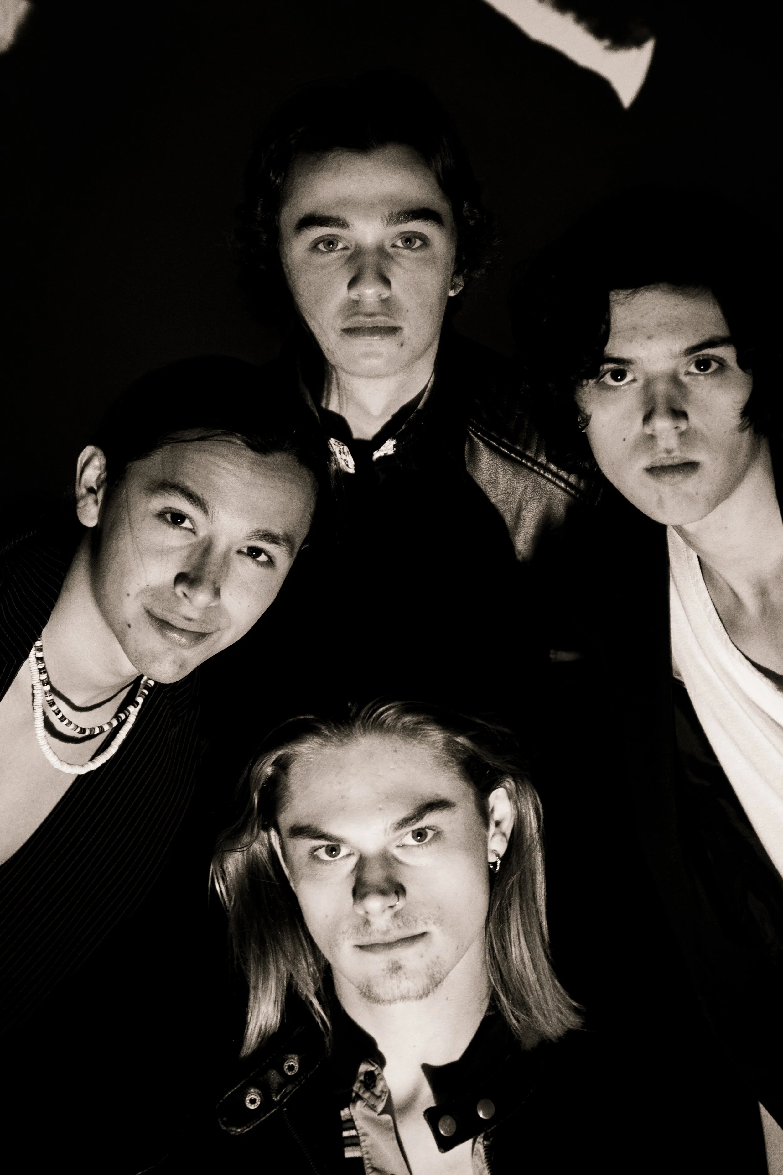 A black and white photo of five young individuals with medium-length hair, some with curly hair, posing closely together for a portrait. They are all looking directly at the camera with neutral or subtle expressions.
