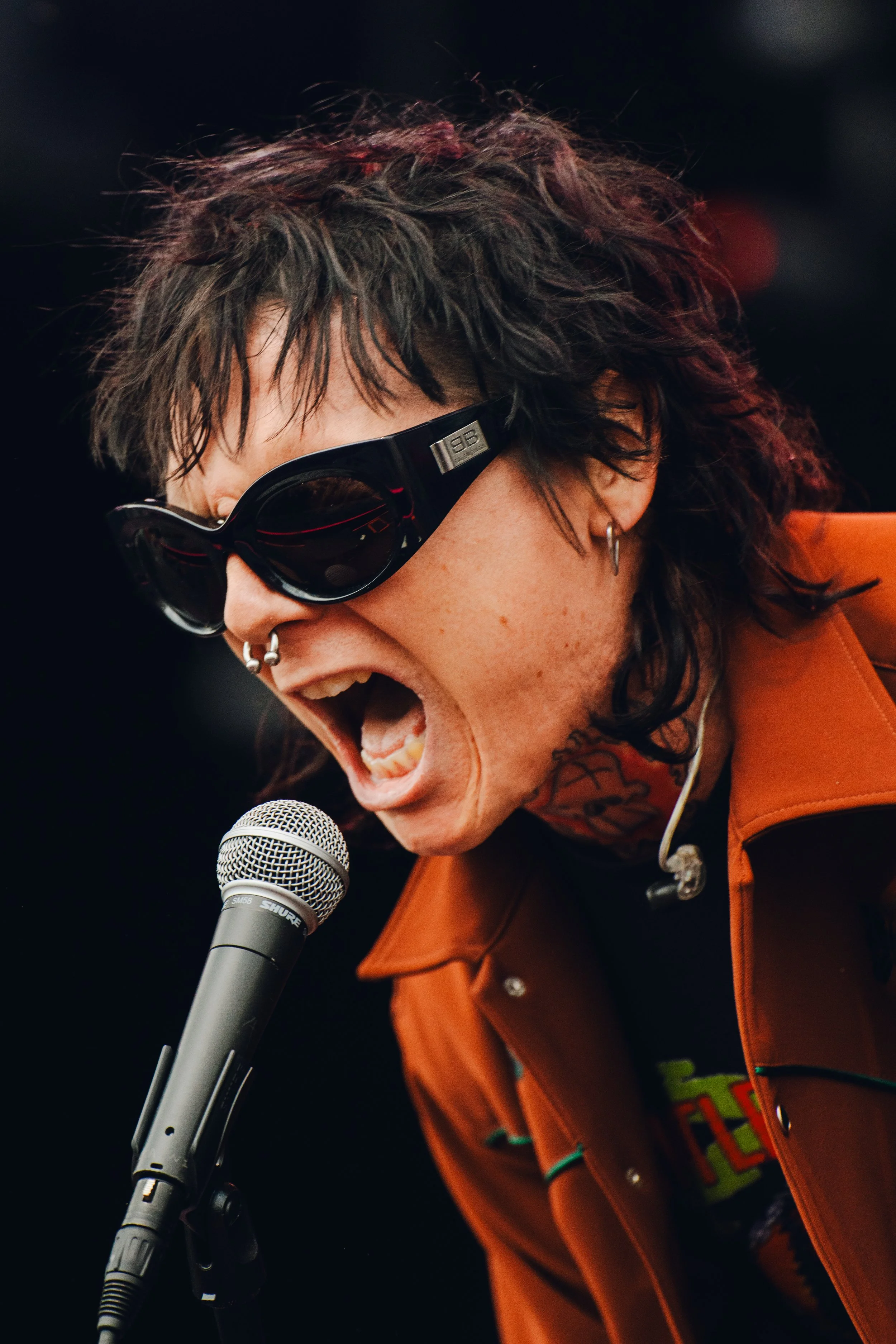 A person with curly hair wearing dark sunglasses and multiple earrings, passionately singing or shouting into a microphone. They are dressed in an orange jacket, with a colorful shirt underneath, and are in a lively setting.