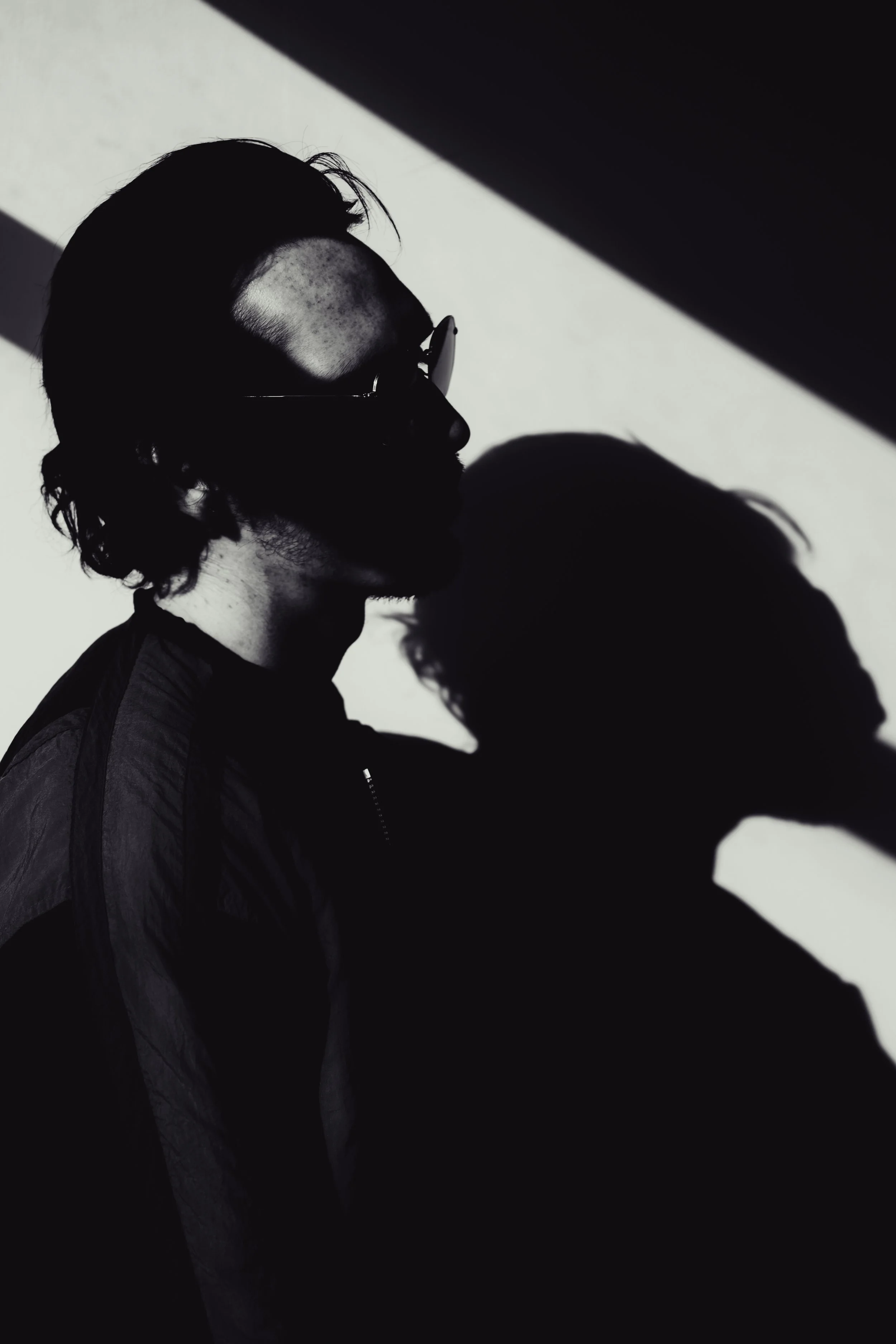 A person with glasses and facial hair, photographed in profile against a black and white background, with shadows creating a high-contrast silhouette.