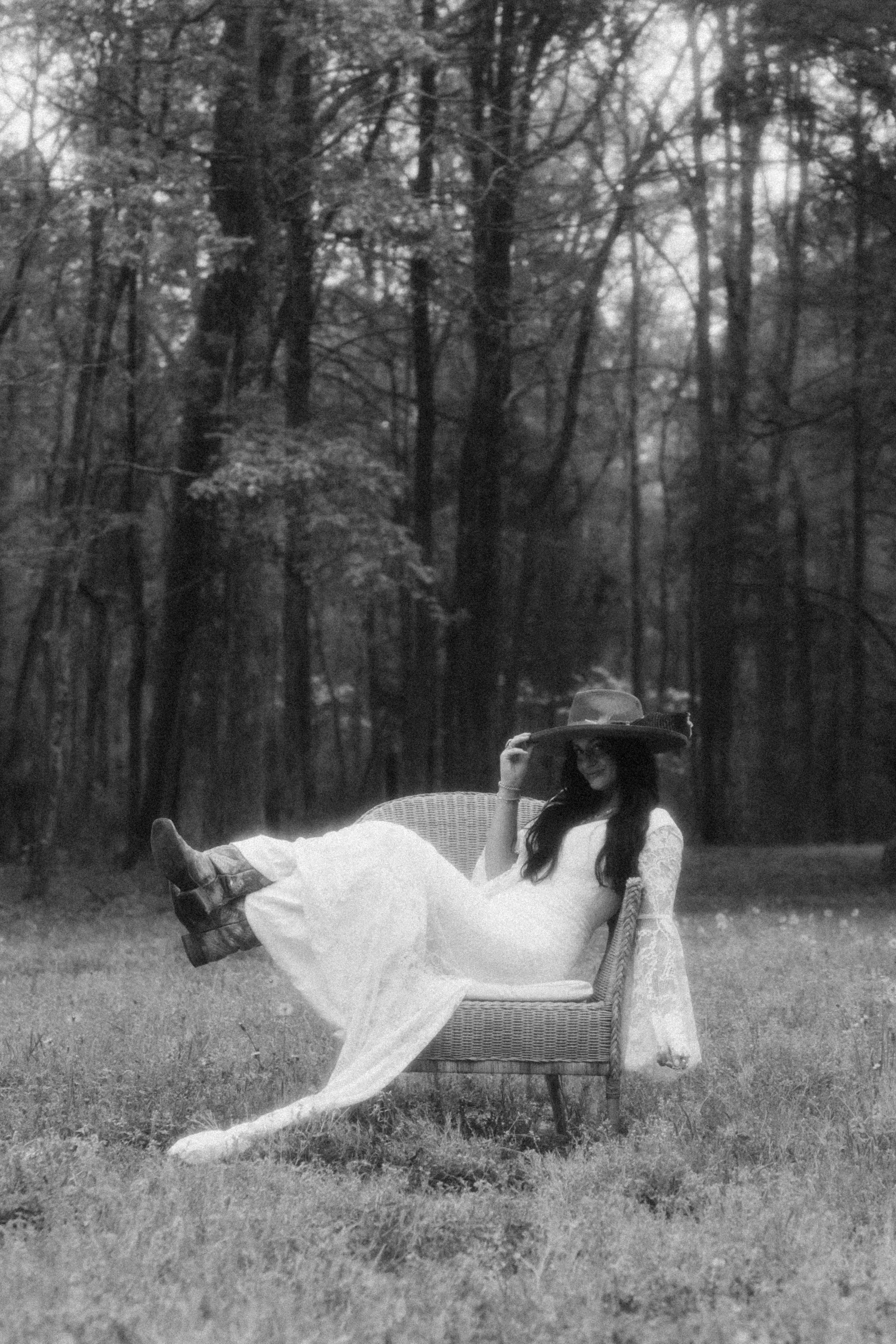 A woman in a white dress and large hat sitting on a wicker chair outdoors in a forested area.