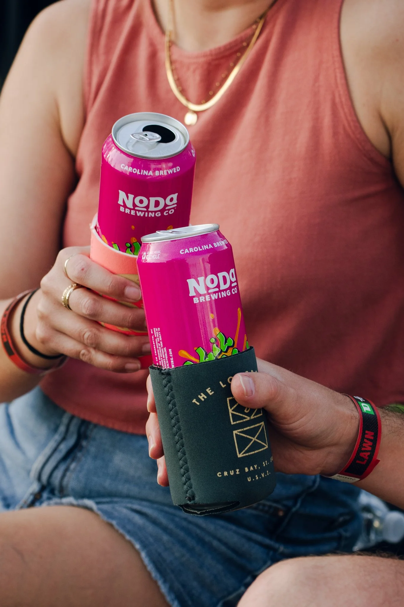 A person wearing a sleeveless pink top and denim shorts holding two cans of Noda Brewing Co drinks in a can holder and a black koozie, with a red and black wristband on their right wrist and a gold ring on their left hand.