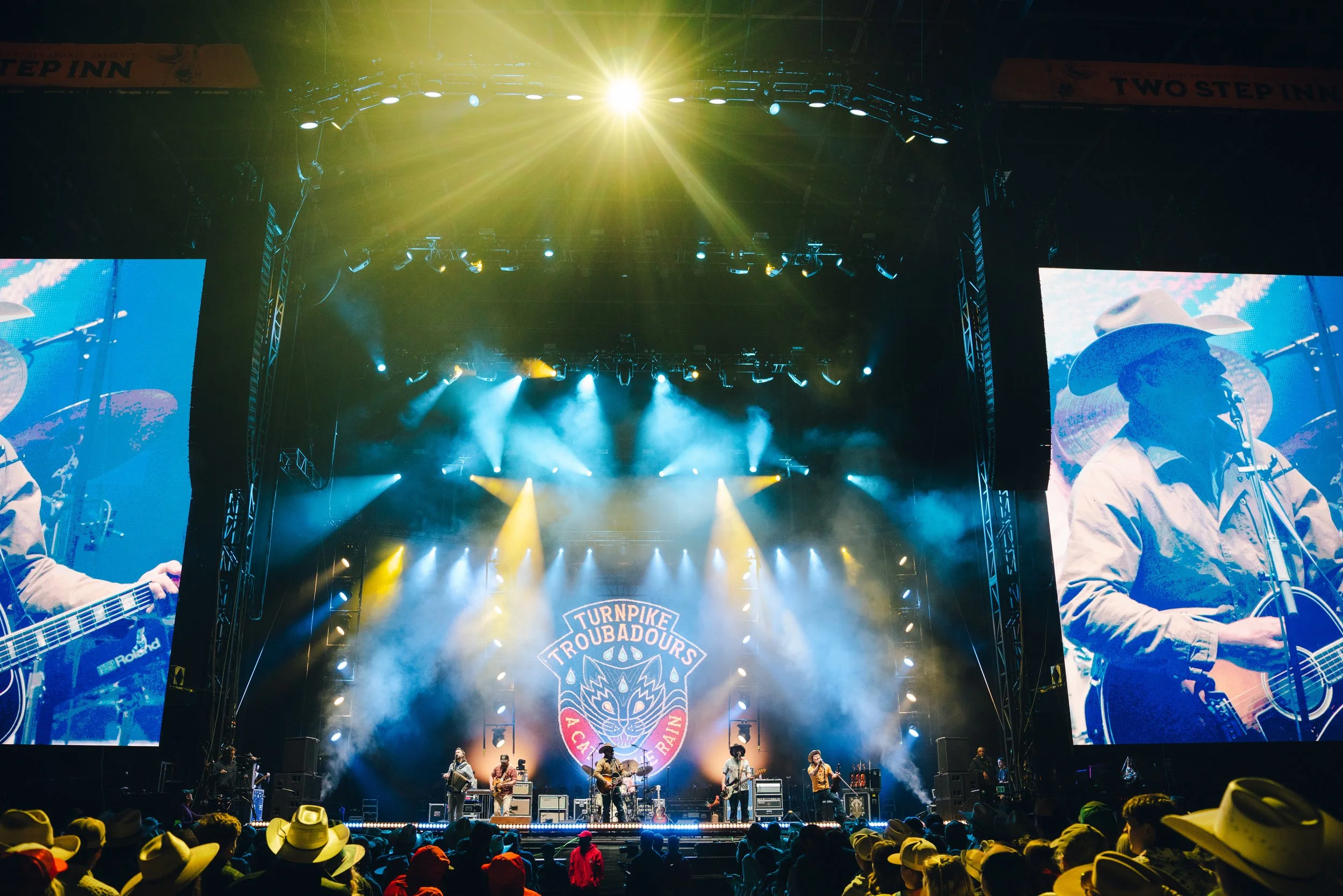 A live outdoor music concert at night with band performing on stage surrounded by bright lights, large screens showing images of band members, and an audience wearing cowboy hats.