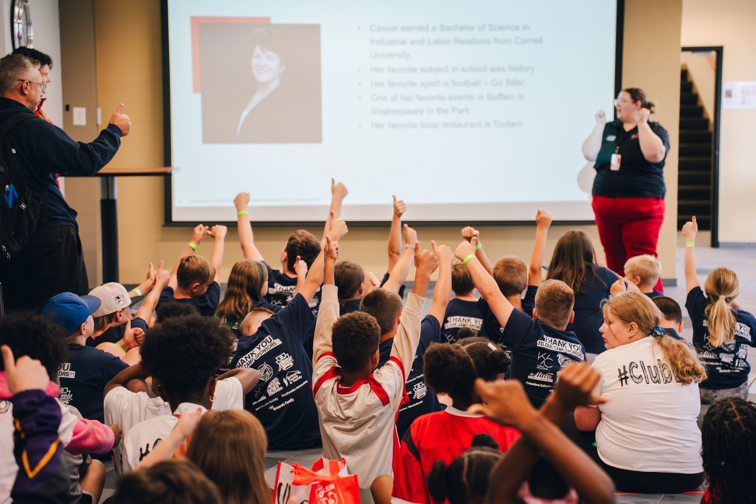 Children sitting on the floor, raising their hands to ask questions or participate, during a presentation or class in a school or conference room. A woman stands at the front, near a large screen displaying a slide with a photo and bullet points. An 