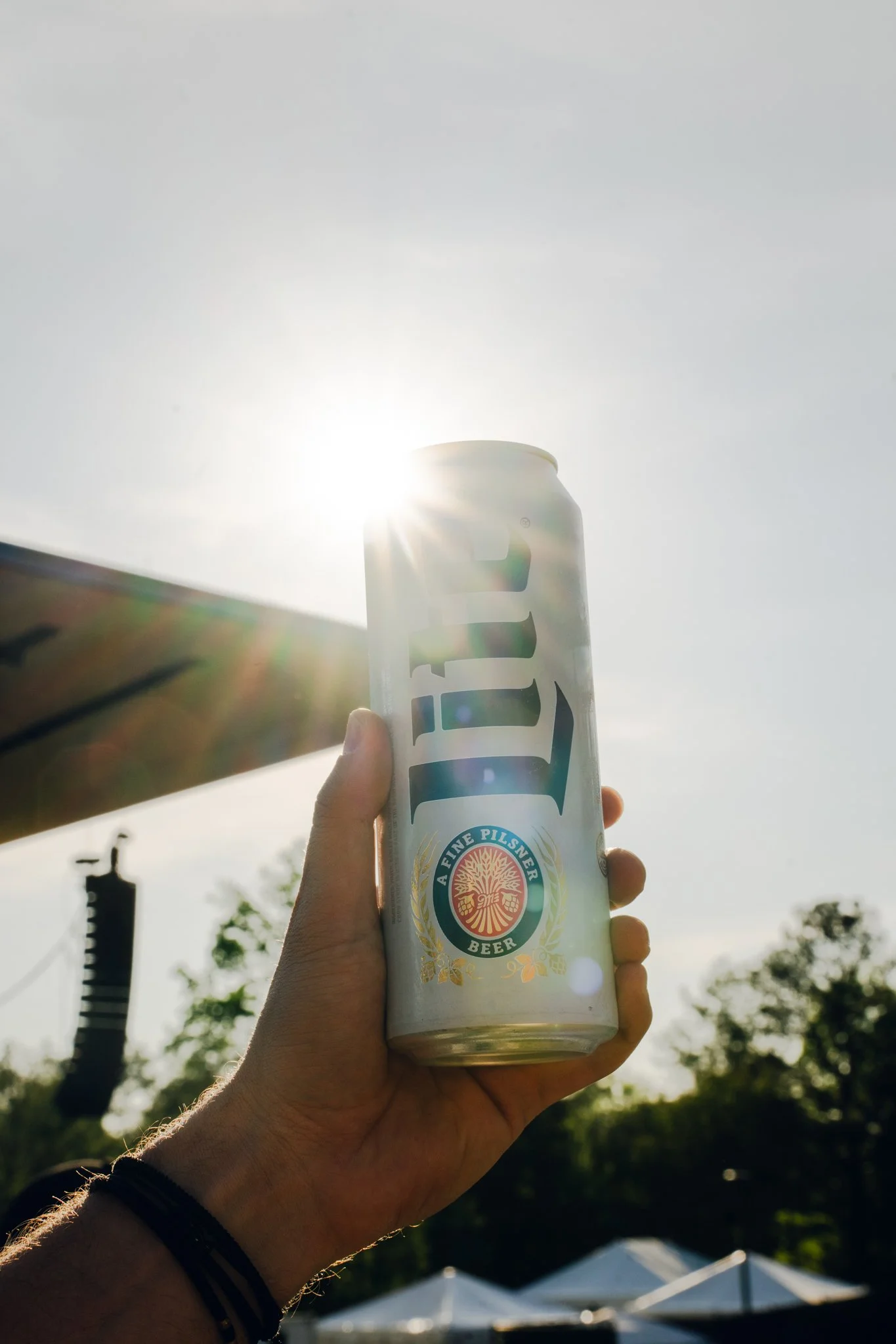 A hand holding a white can of beer with a label reading 'A Fine Pilsner Beer,' with the sun shining brightly behind the can against a clear sky, with some outdoor tents and trees in the background.