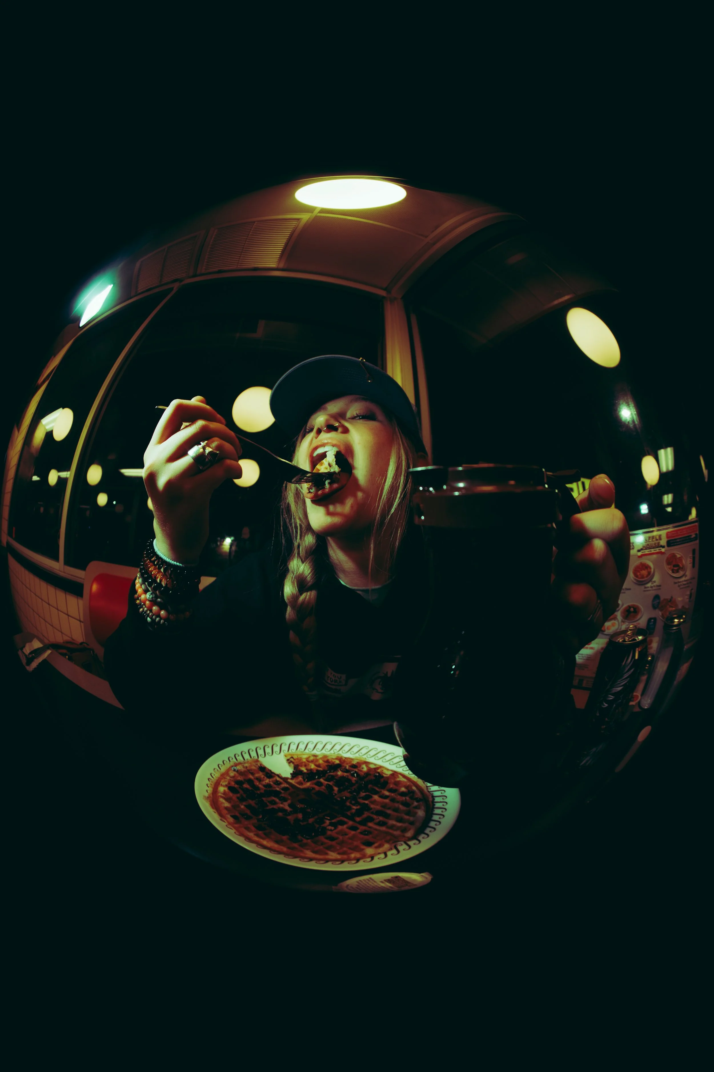 A woman with long blonde hair wearing a baseball cap and multiple bracelets, sitting in a restaurant, eating a slice of pizza, with a jar of soda in front of her, and a plate of waffles on the table. The restaurant has circular lights reflected in th