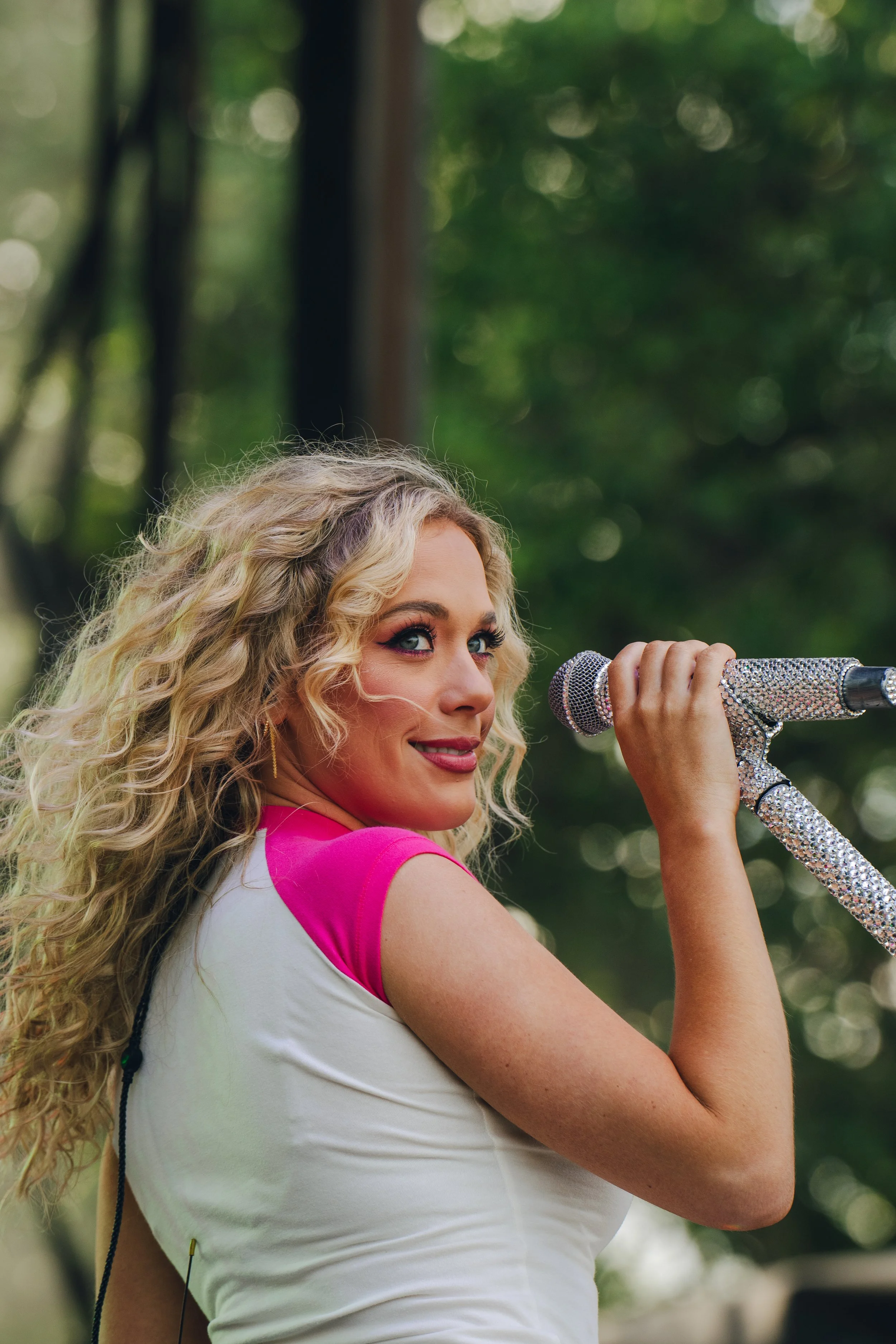 A woman with curly blonde hair holding a sparkling microphone and smiling outdoors, wearing a white and pink sports top.