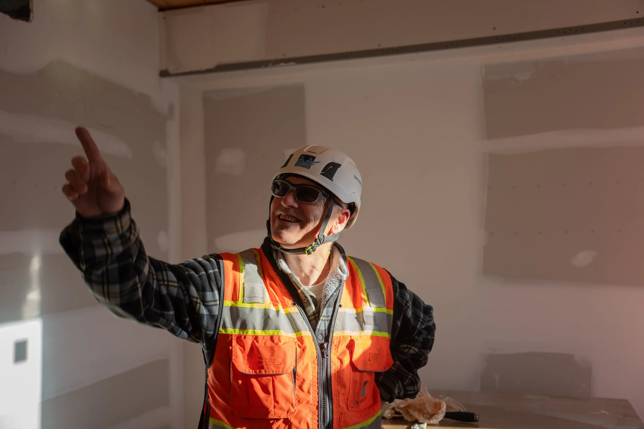 A man wearing a construction helmet, safety glasses, and an orange safety vest pointing upwards while standing inside a building under construction.