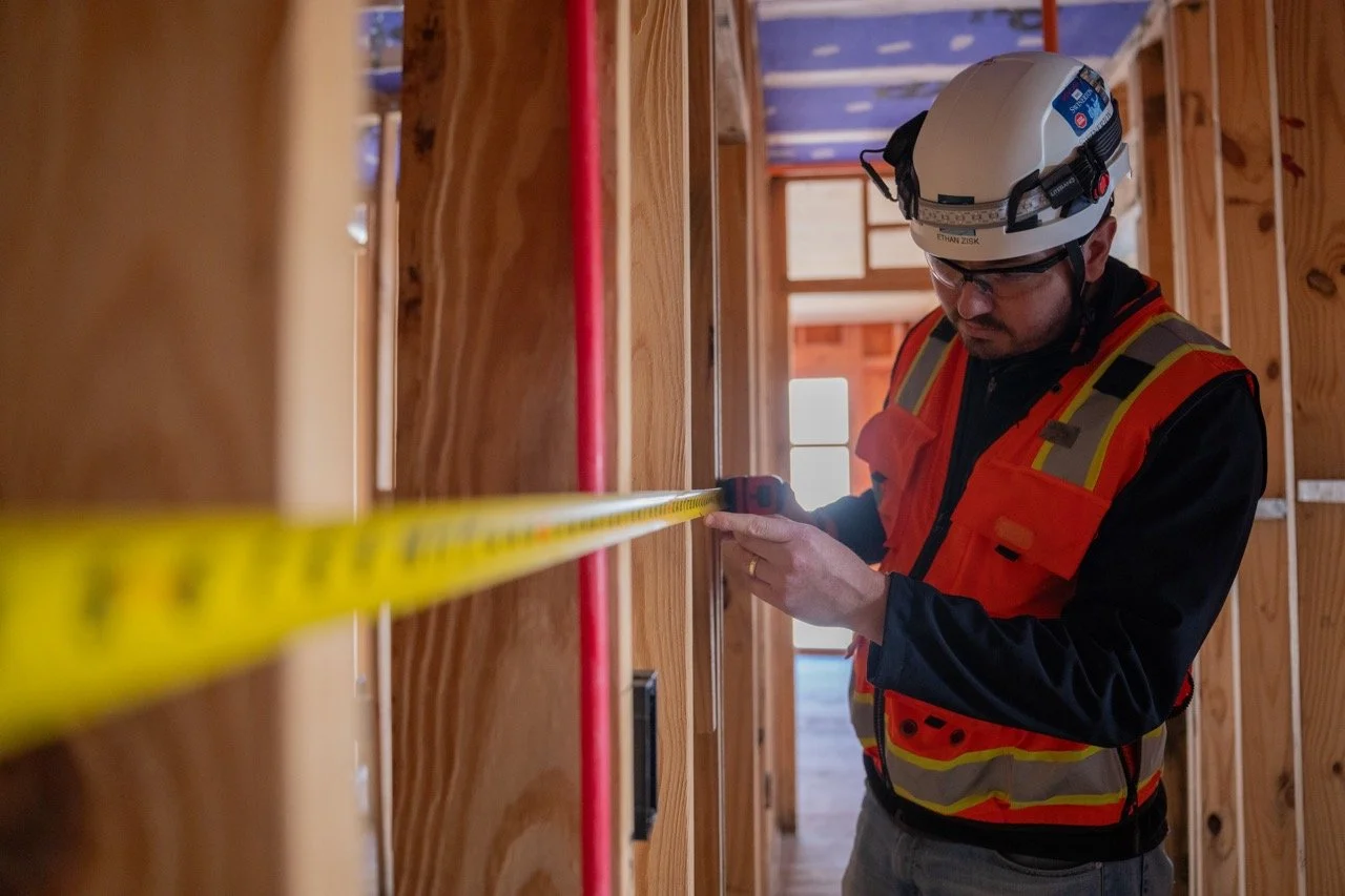 A construction worker in safety gear measuring a wooden wall using a tape measure inside a building under construction.