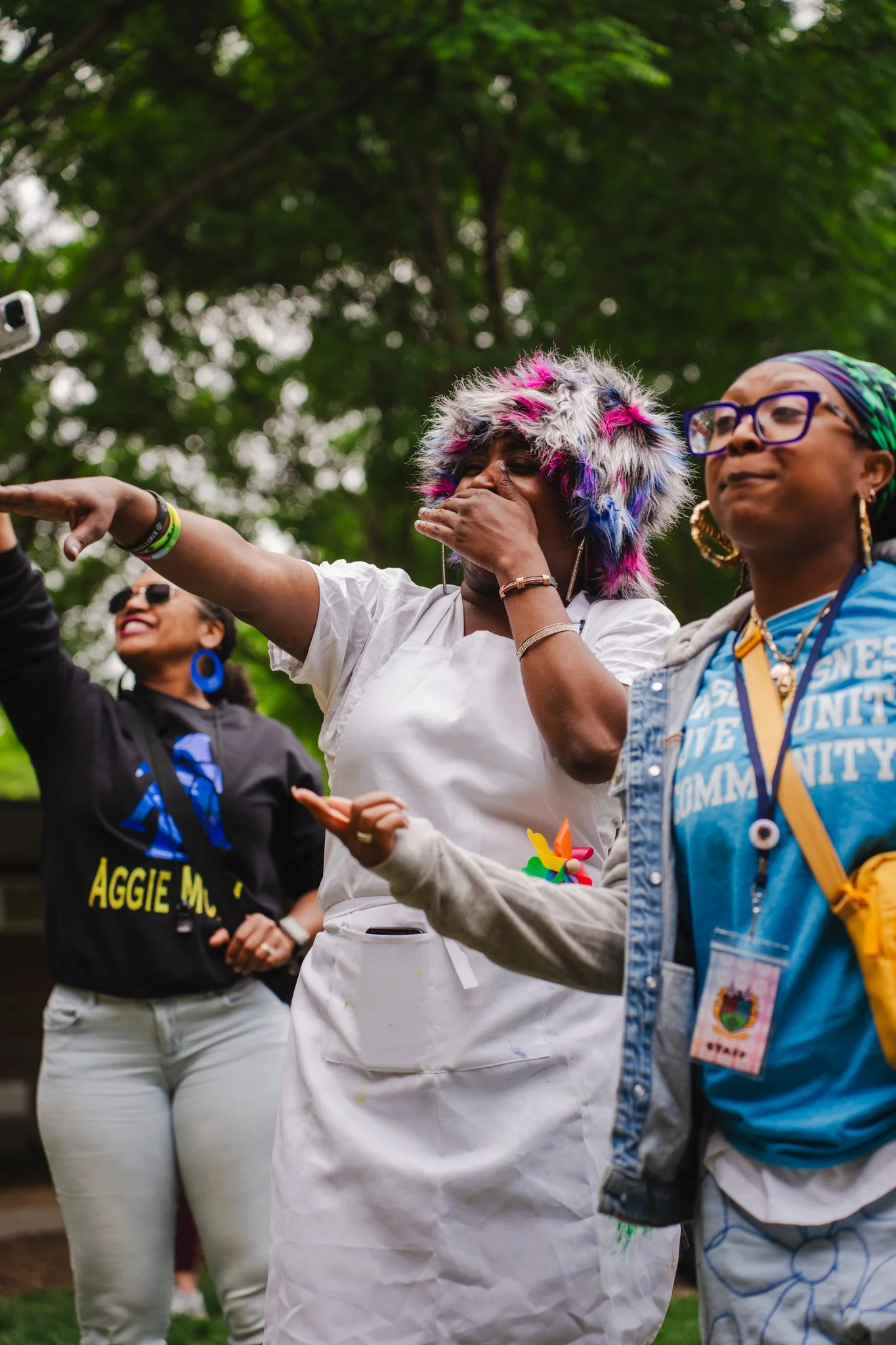 Group of women at an outdoor event, one wearing a colorful furry hat and another with glasses and a headscarf, engaging in cheerful conversation.
