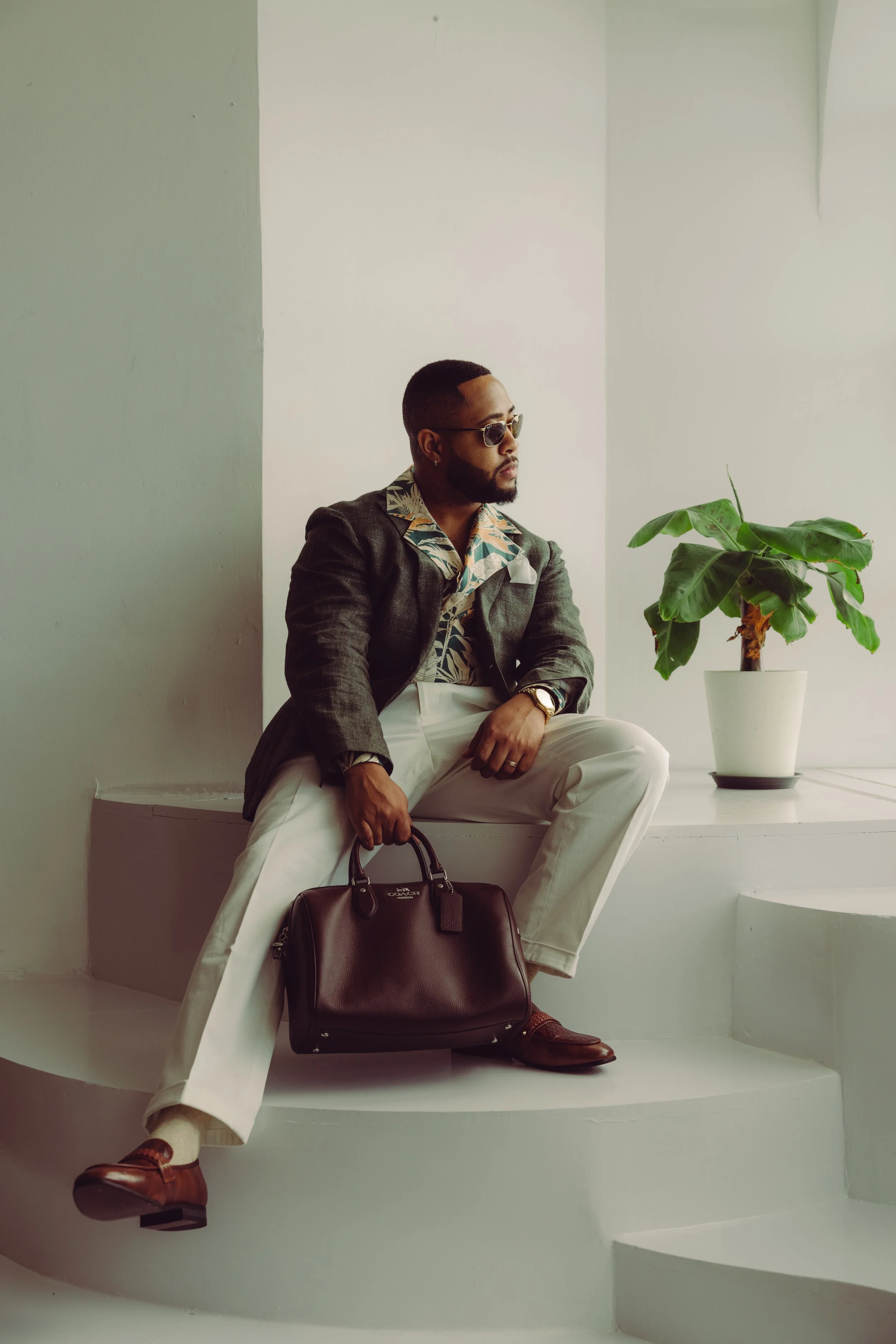 A man sitting on a white step, wearing sunglasses, a suit jacket, beige pants, and brown shoes. He is holding a brown handbag, with a potted plant with large green leaves next to him and a plain white wall in the background.