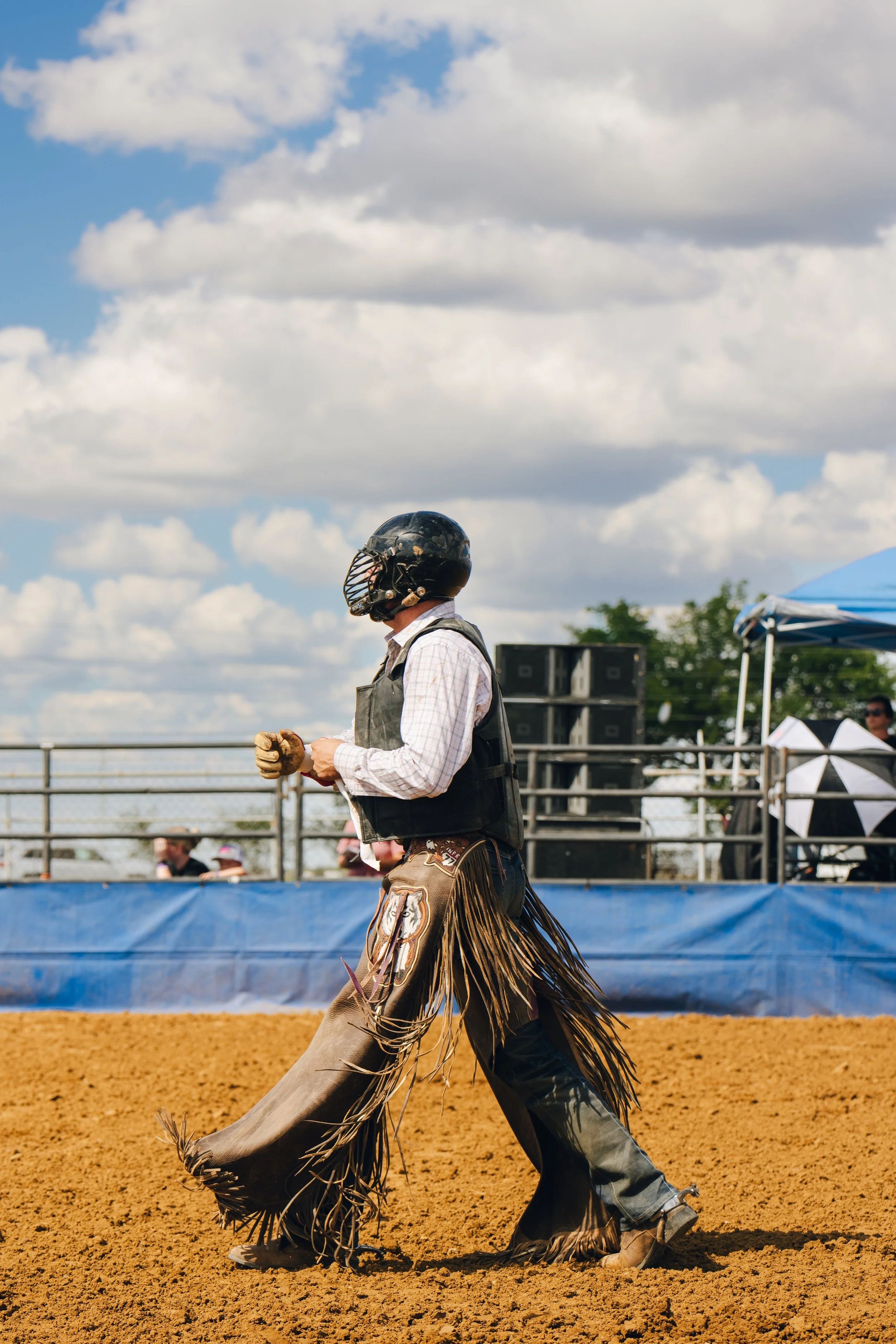 Person wearing a black helmet, plaid shirt, and fringed chaps riding a horse on a dirt track during an outdoor rodeo event under a partly cloudy sky.