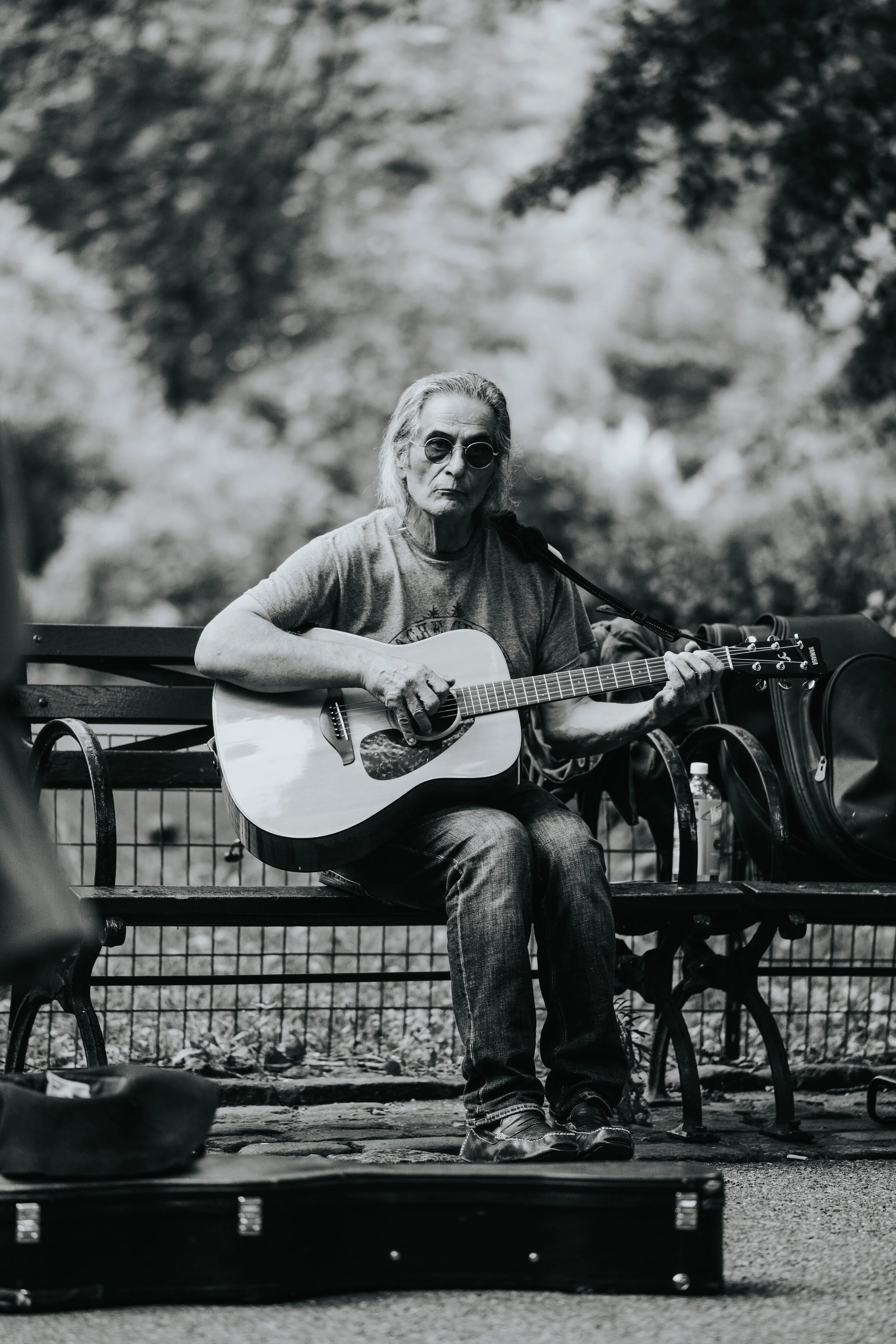 An elderly man with long gray hair and wearing sunglasses, a T-shirt, and jeans, sitting on a park bench playing an acoustic guitar.