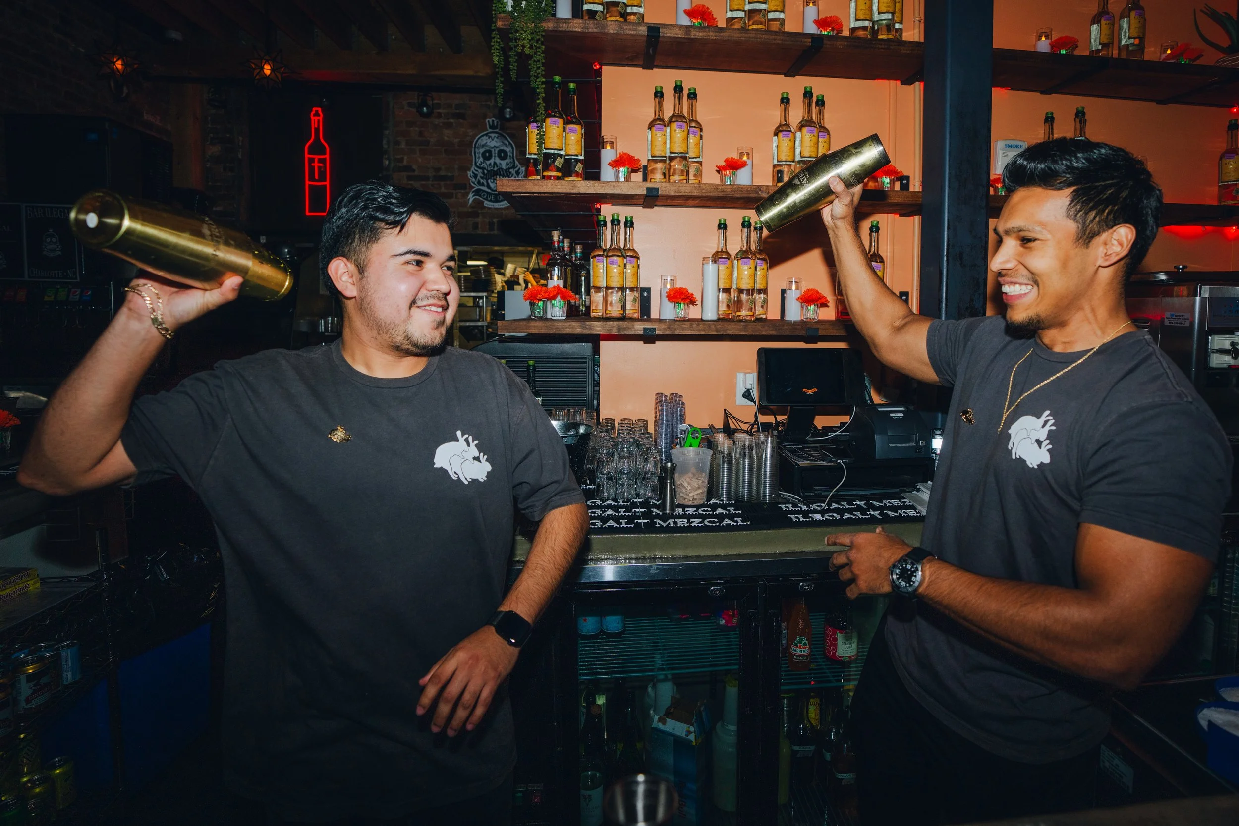 Two male bartenders smiling and throwing metal shakers in the air inside a bar. The background shows shelves with bottles and decorative flowers.