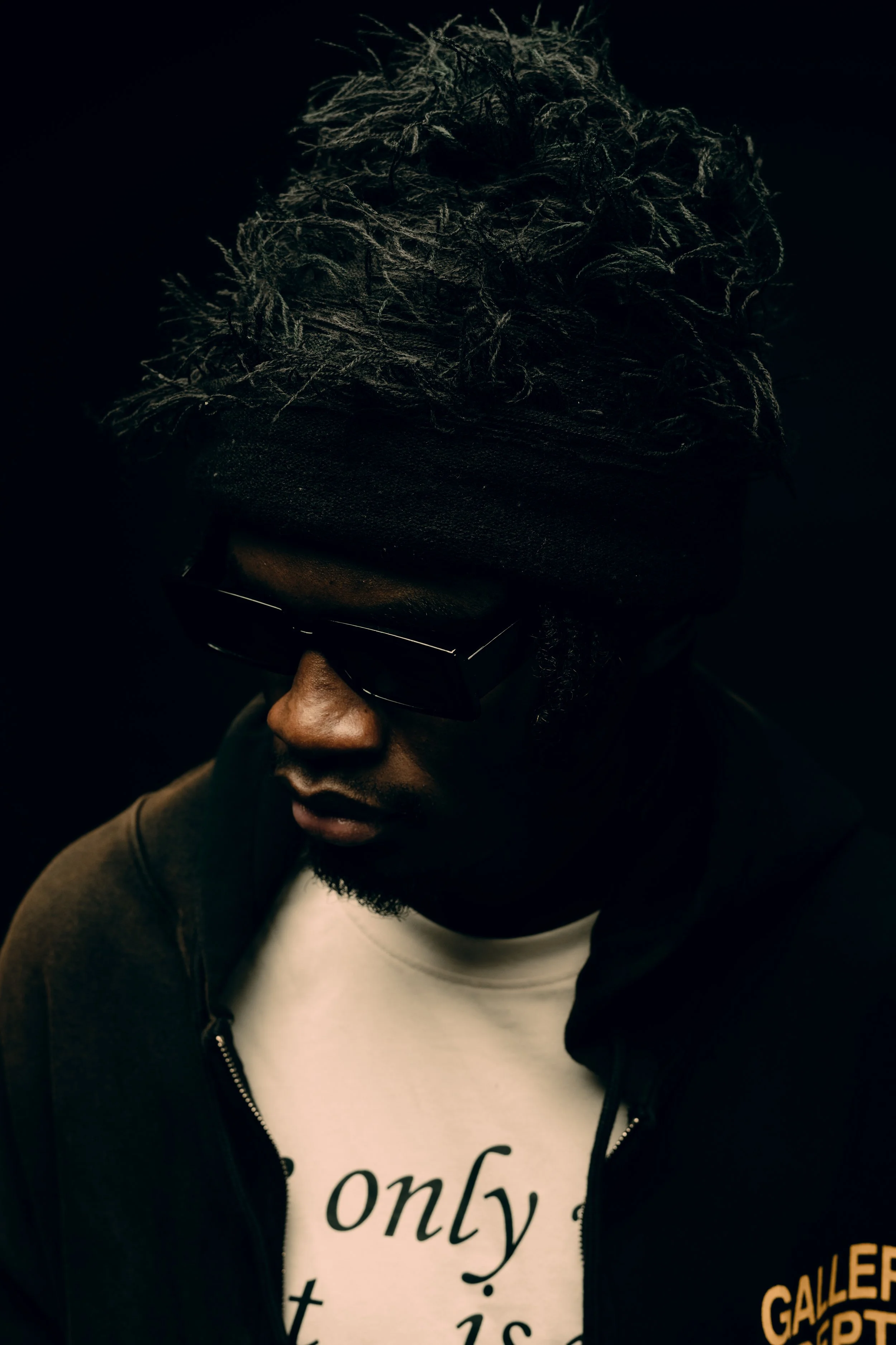 A person wearing black rectangular sunglasses, a white t-shirt with black text that reads "only" and additional text that is not fully visible, a dark zip-up hoodie with yellow lettering that says "GALLERY", and a large black hat with textured, tangl