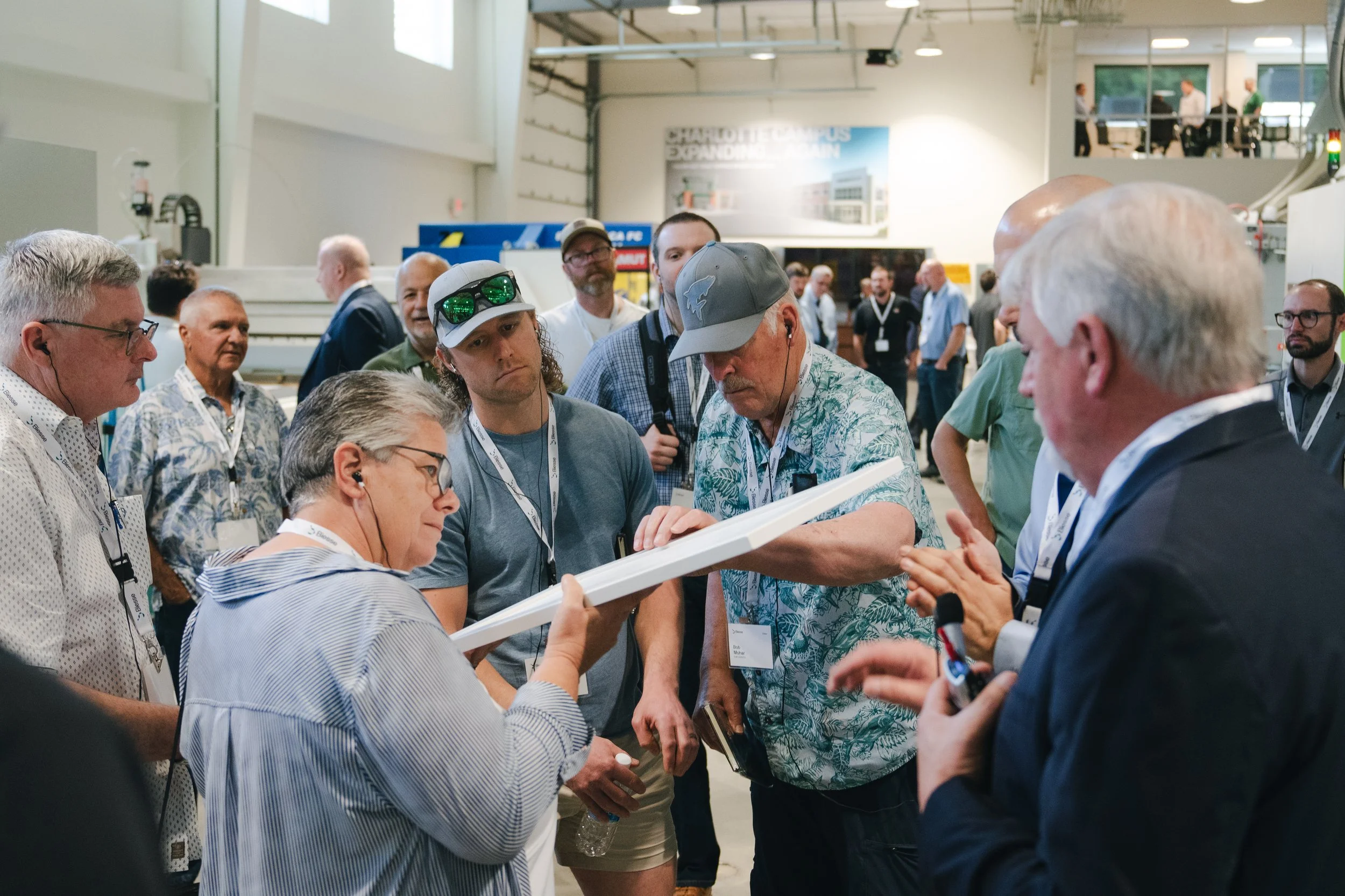 A group of people gathered in an exhibition hall, engaged in a discussion, with some looking at a tablet device being held by a woman.