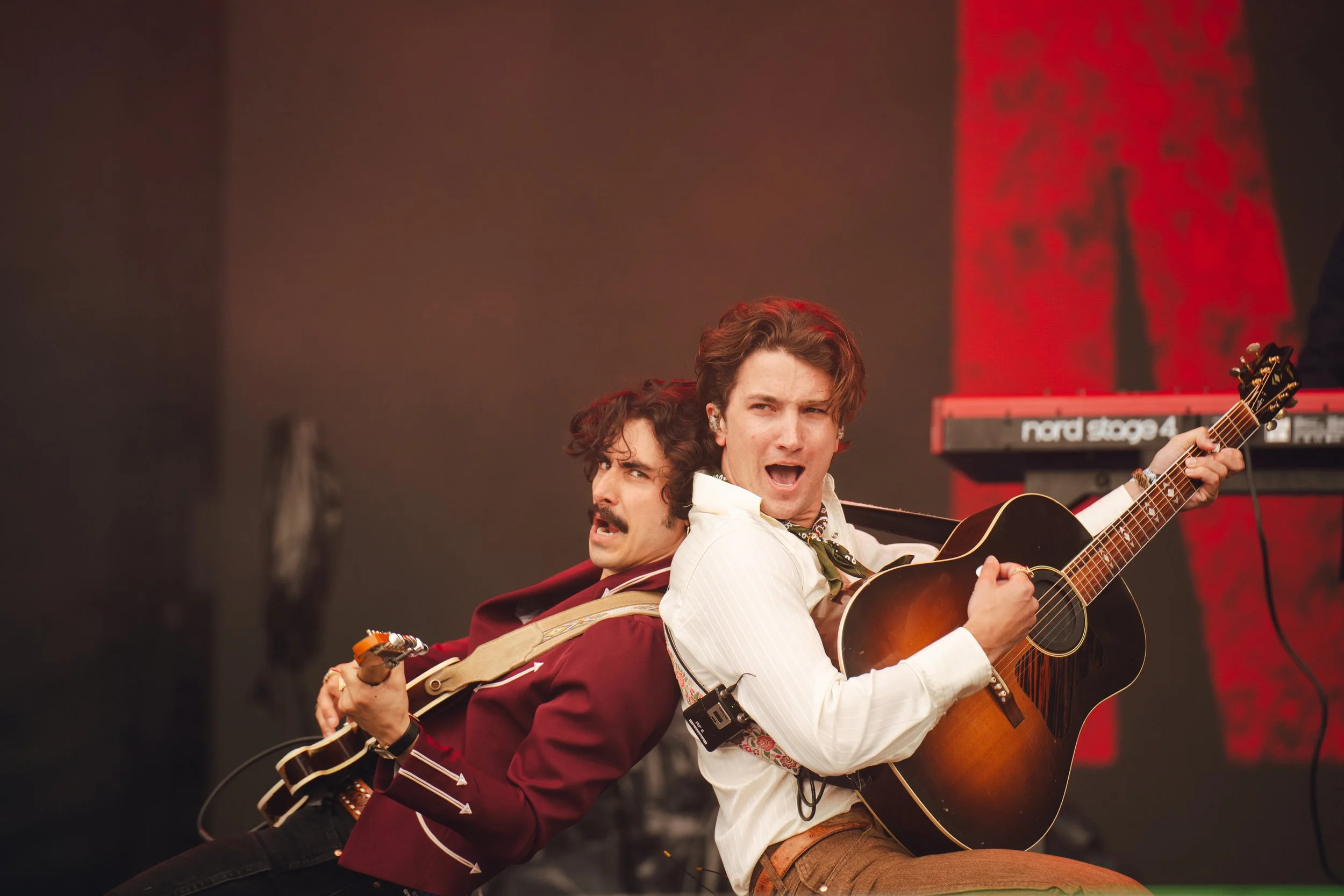 Two men performing on stage with guitars, one singing, in front of a red and black background.