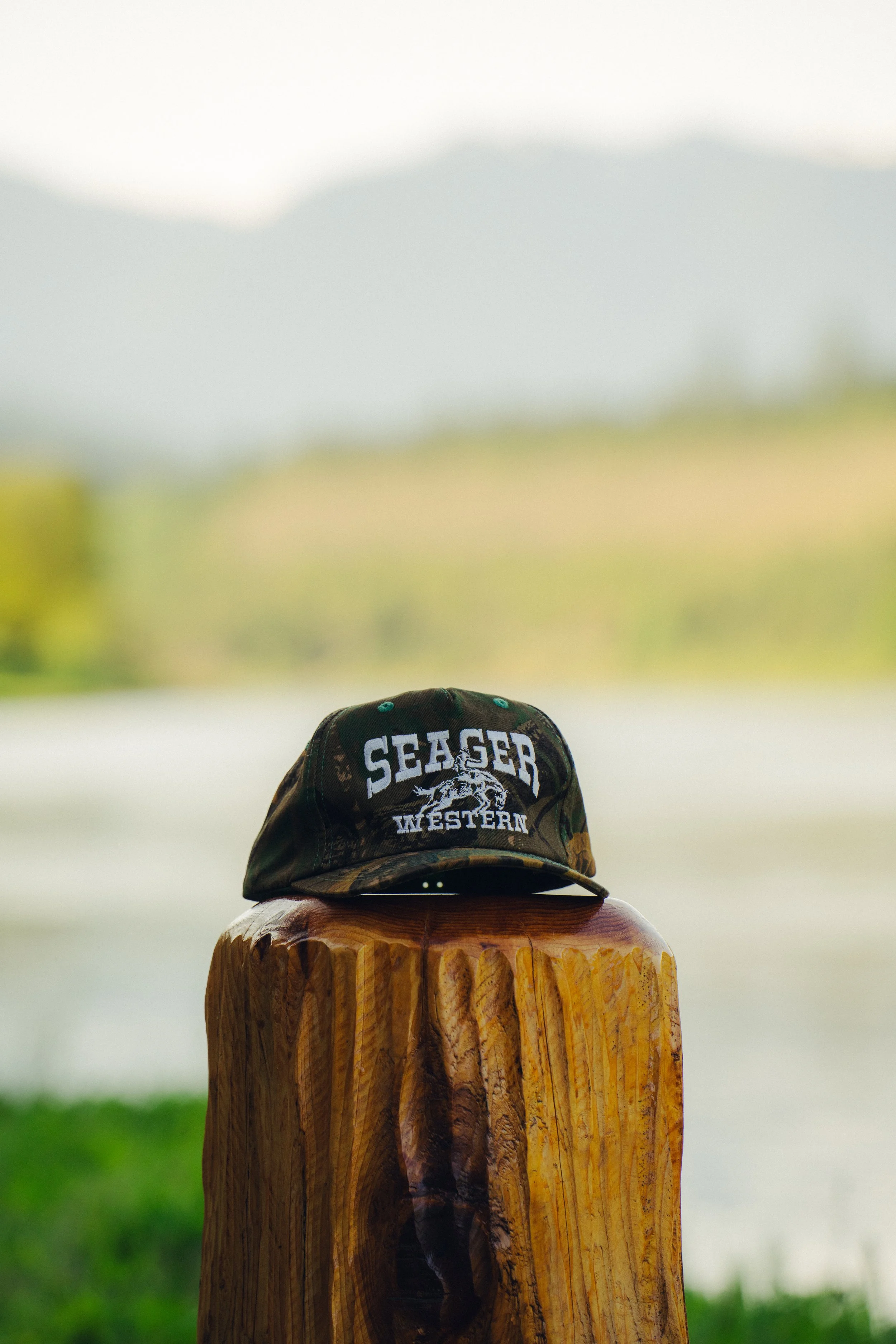 A camouflage baseball cap with the words 'SEAGER WESTERN' and an image of a horse, placed on a wooden post outdoors with a blurred grassy field and cloudy sky in the background.