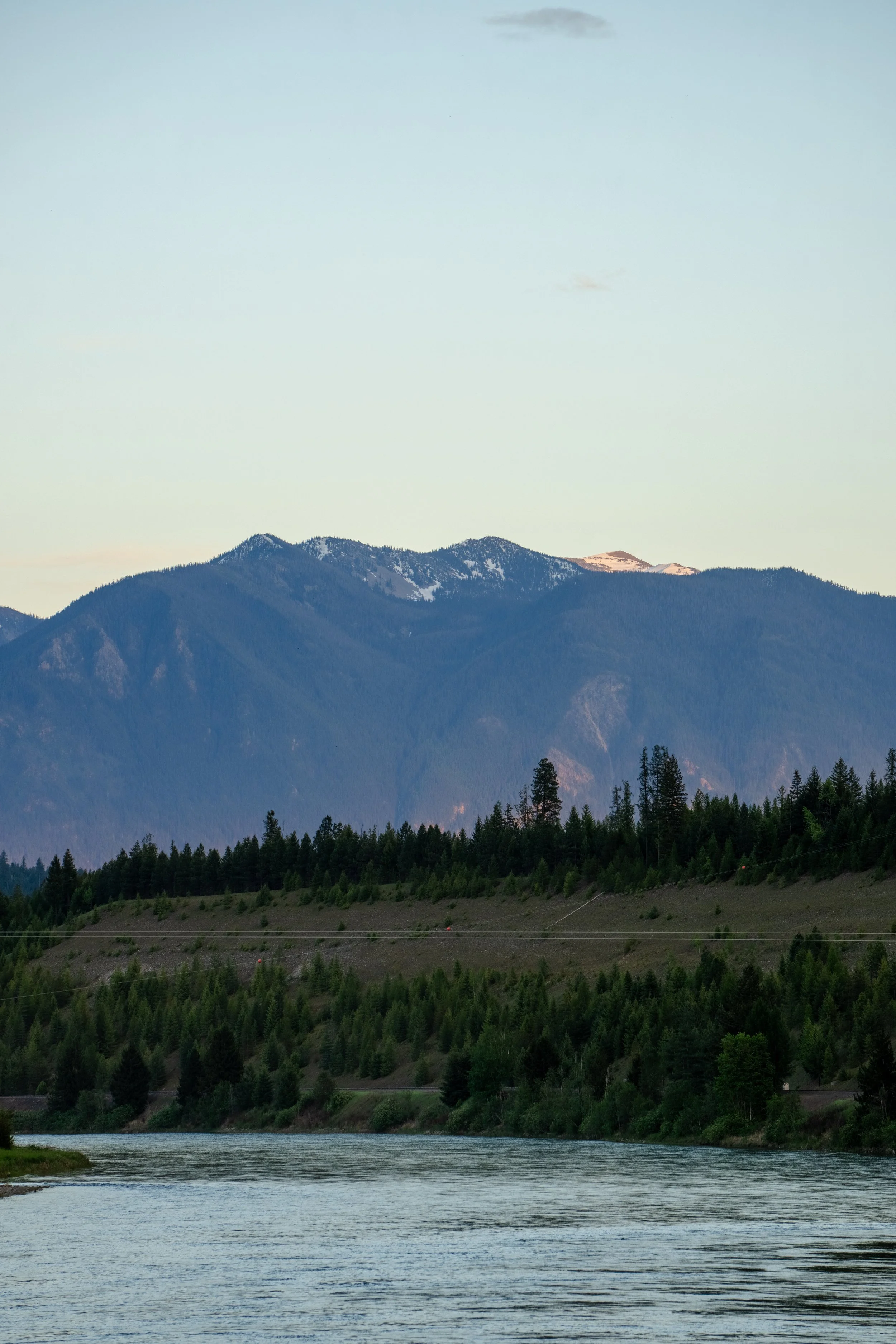 Scenic view of a mountain range with snow patches, a forested hillside, and a body of water in the foreground under a partly cloudy sky.