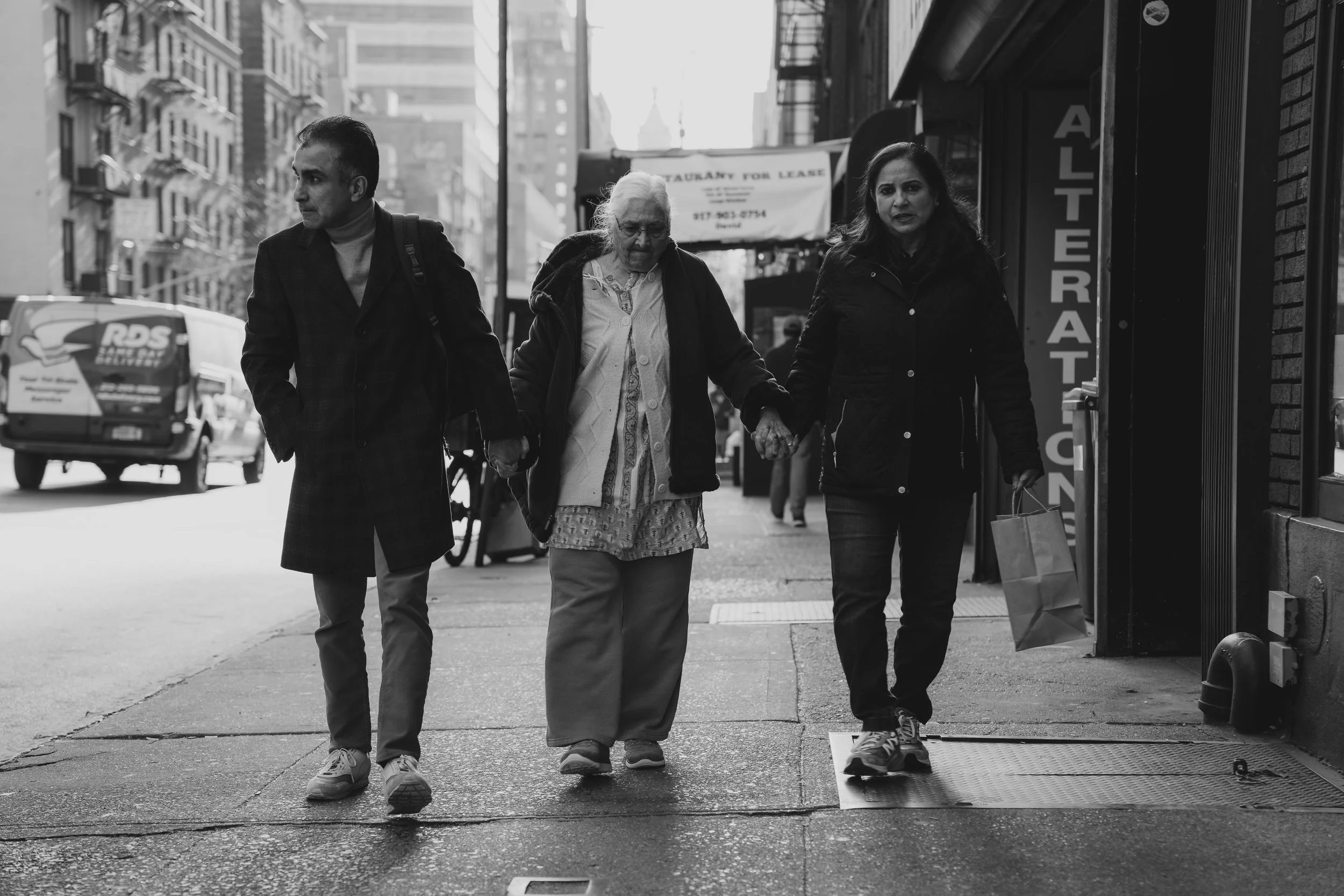Three people walking on a city sidewalk, holding hands. The elderly woman is in the middle, with a man on her left and a woman on her right. The woman is carrying shopping bags. The background shows tall buildings, a parked van, and a storefront sign