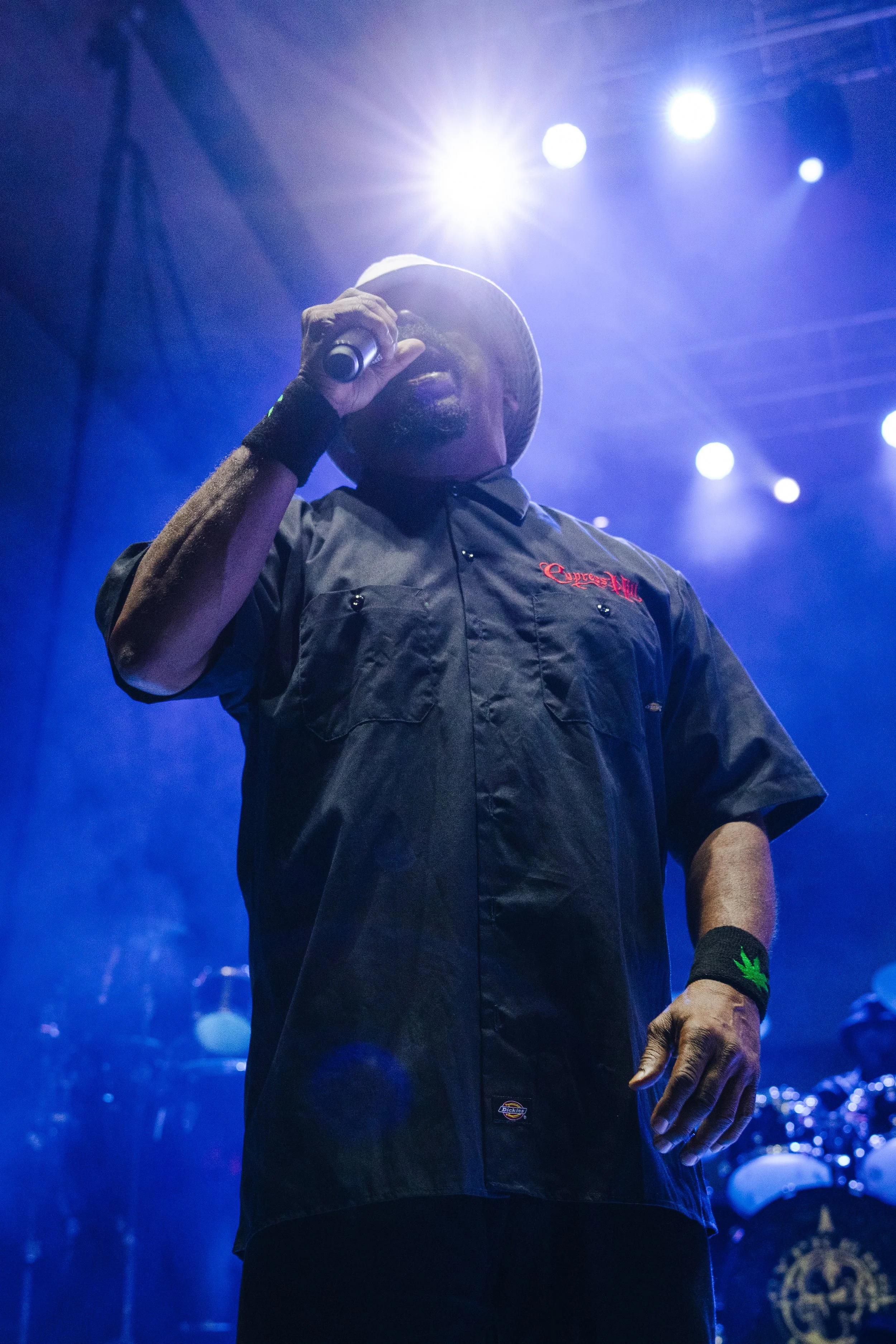 A man wearing a baseball cap and black contractor shirt with red embroidery is performing on stage, holding a microphone to his mouth, illuminated by purple and white stage lights.