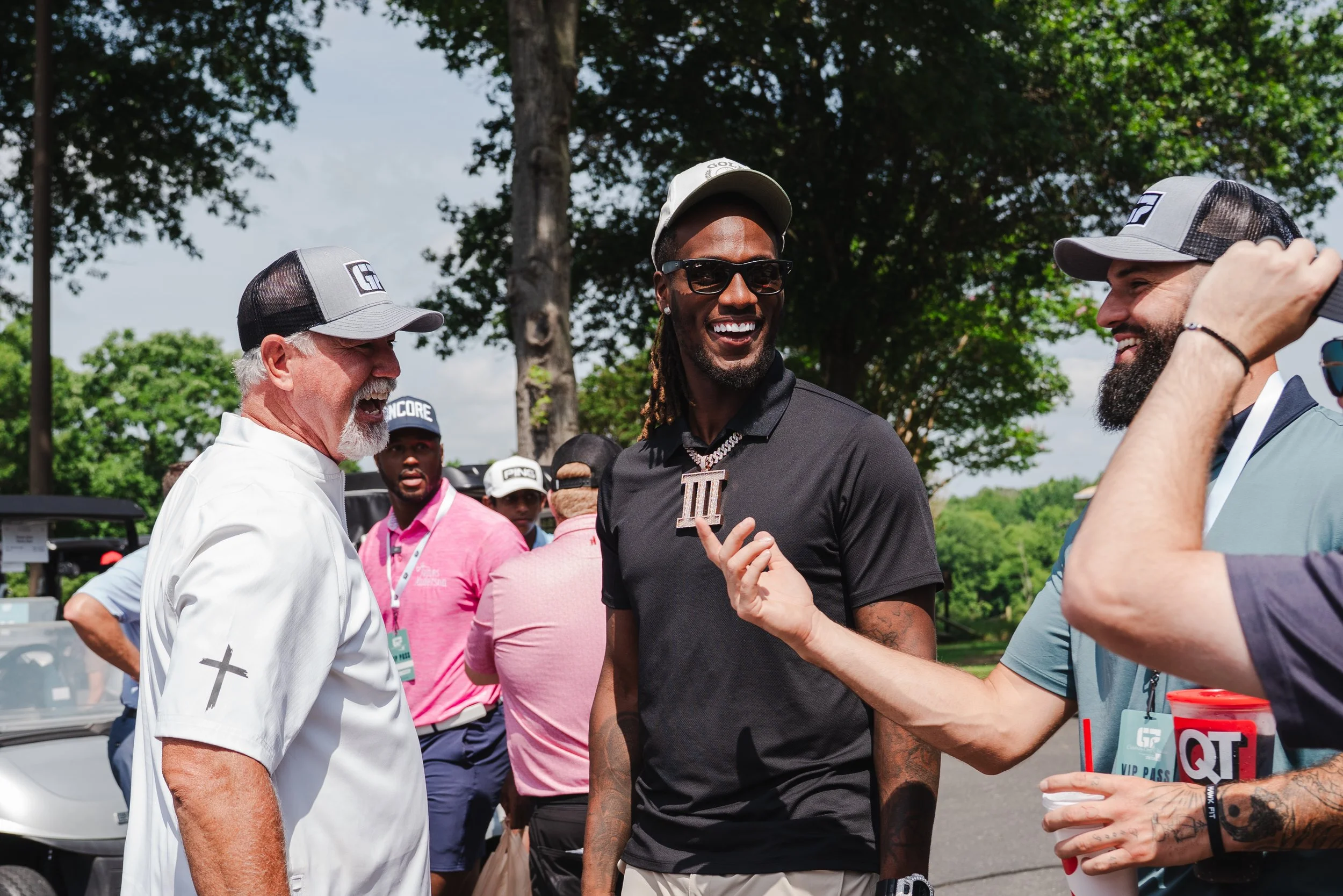 Group of men, including a man in a white baseball cap and a man in a black shirt with a large pendant, engaging in conversation outdoors during a sunny day with trees in the background.