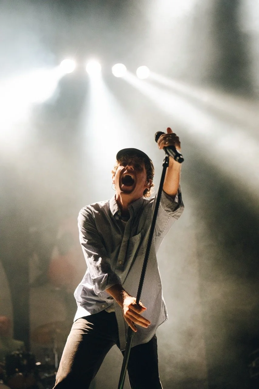 A male singer passionately performing on stage, holding a microphone stand, with bright stage lights shining down from above.
