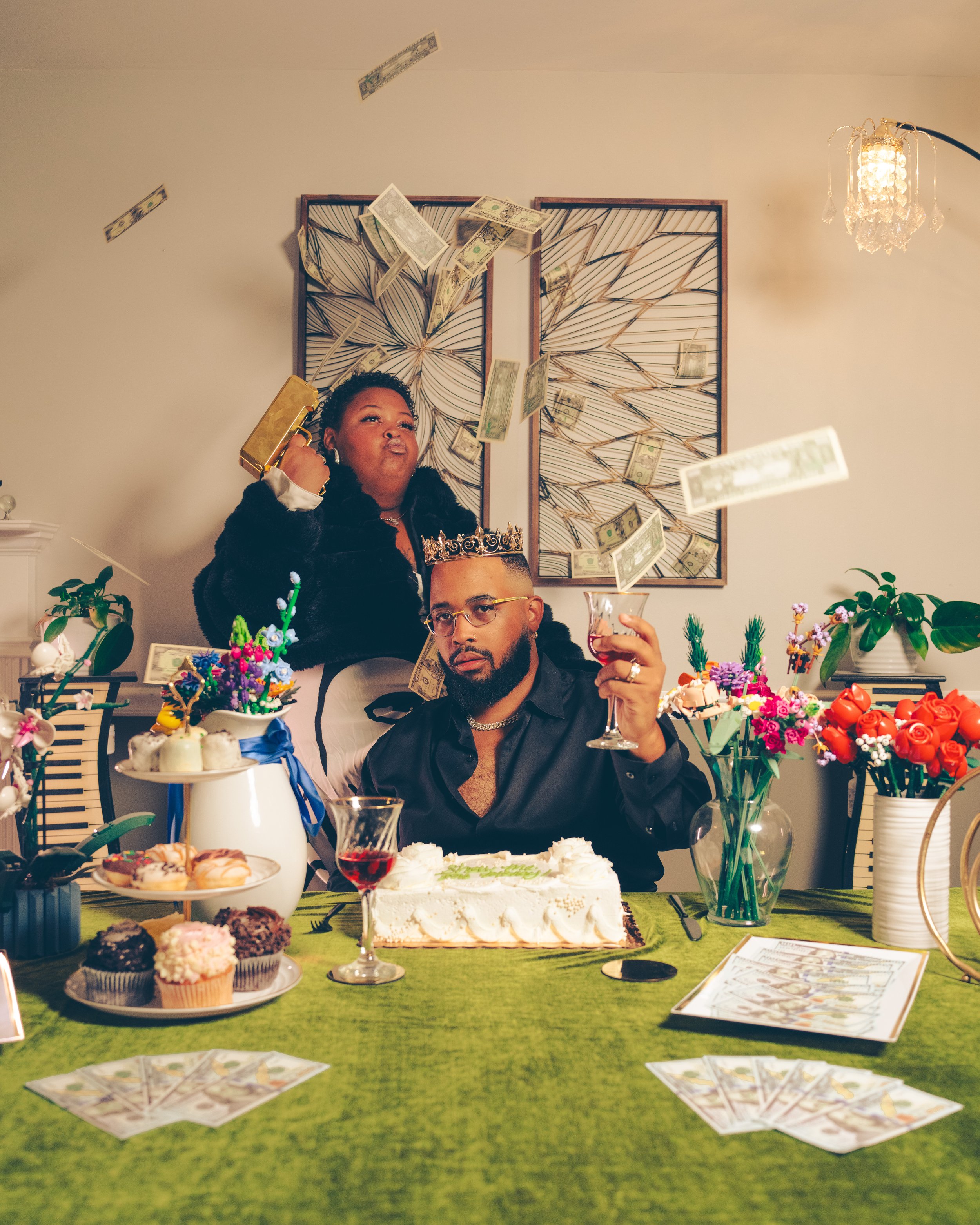 A woman and a man at a celebration with a cake, cupcakes, and game cards on the table, surrounded by flowers and decorations, with money falling from above.