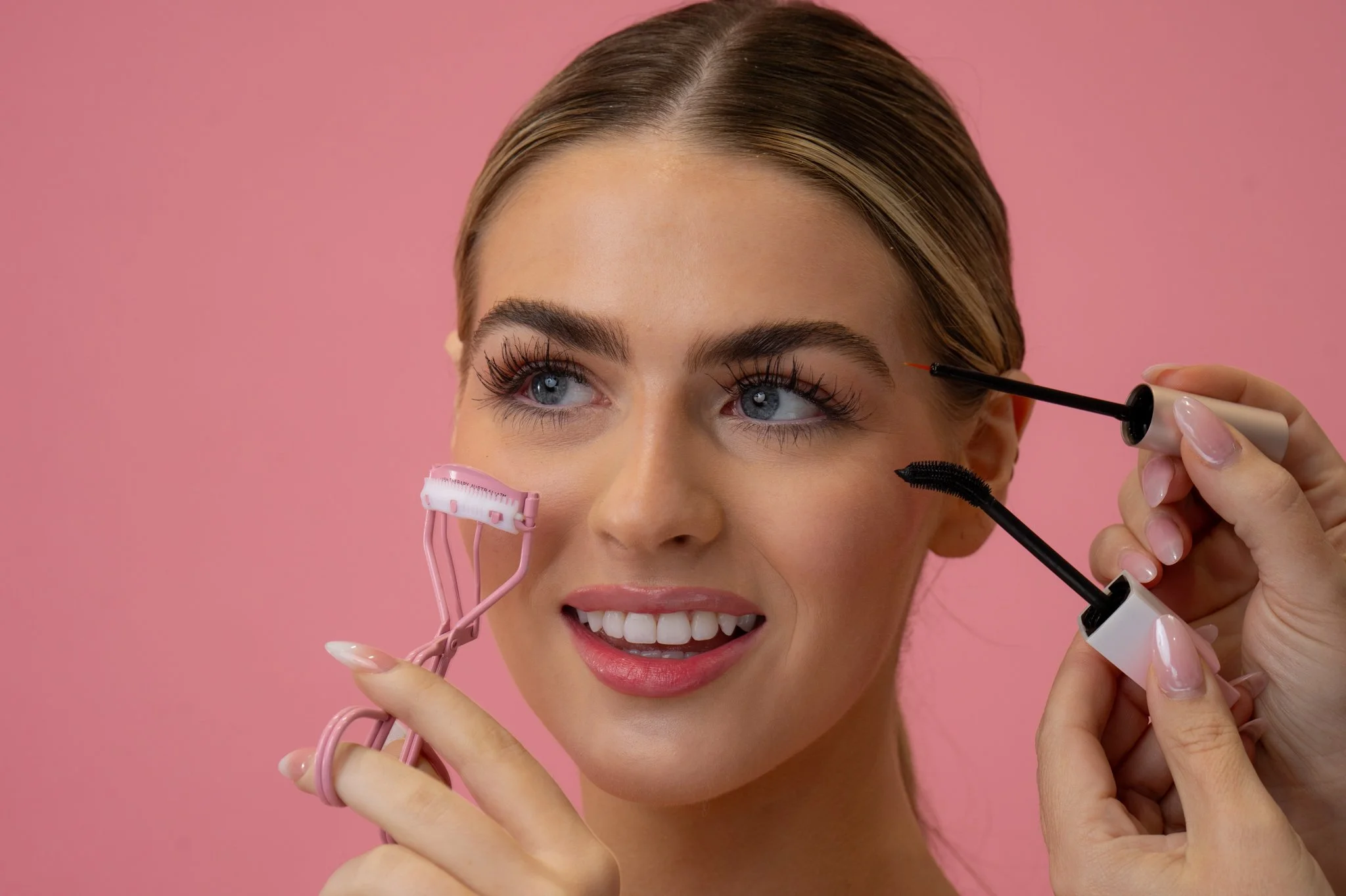 A woman with blue eyes and brown hair is smiling while applying makeup using makeup tools against a pink background.