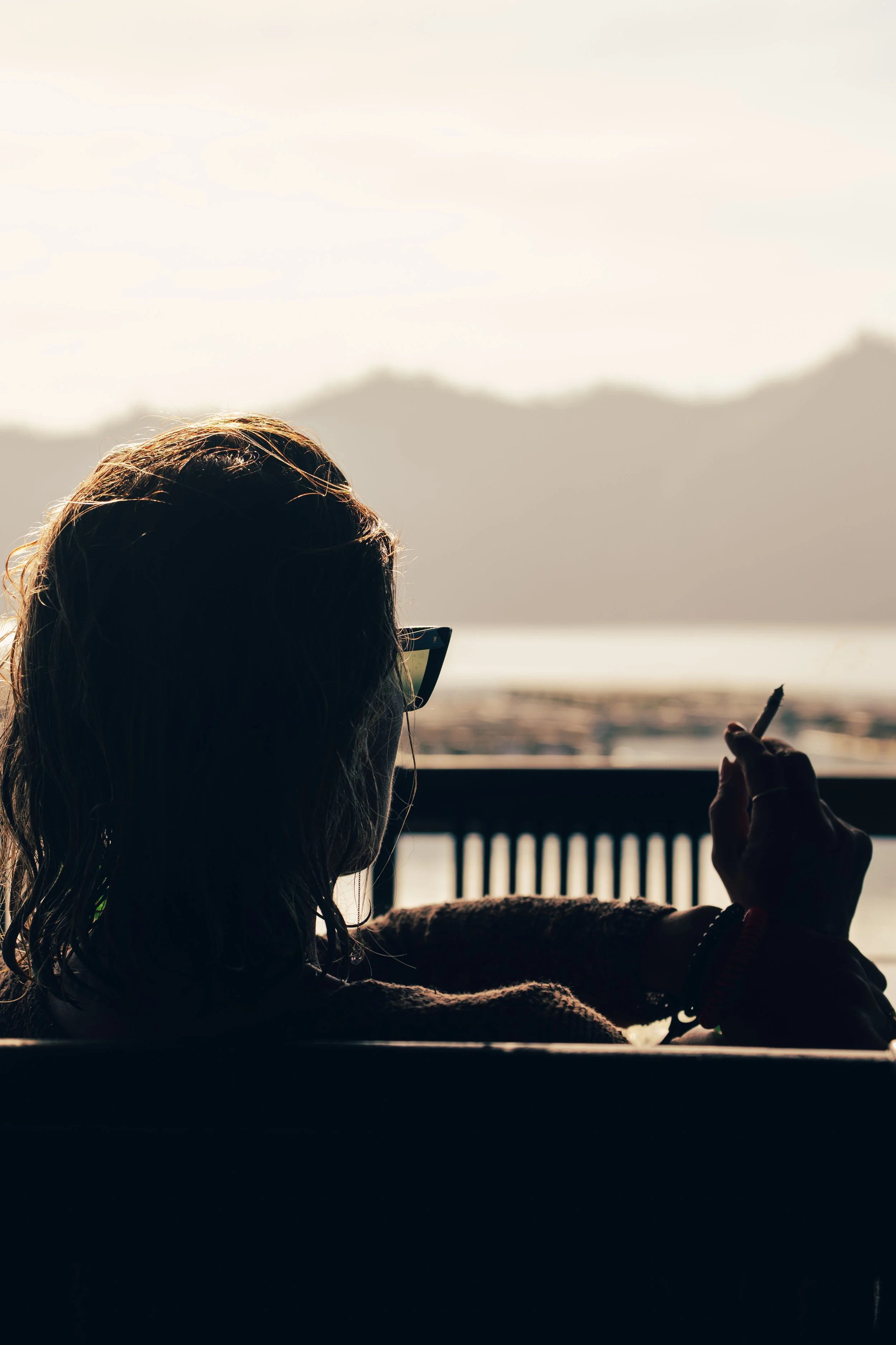 A person with long hair and glasses sitting outside, looking at a body of water with mountains in the background and the sky above.
