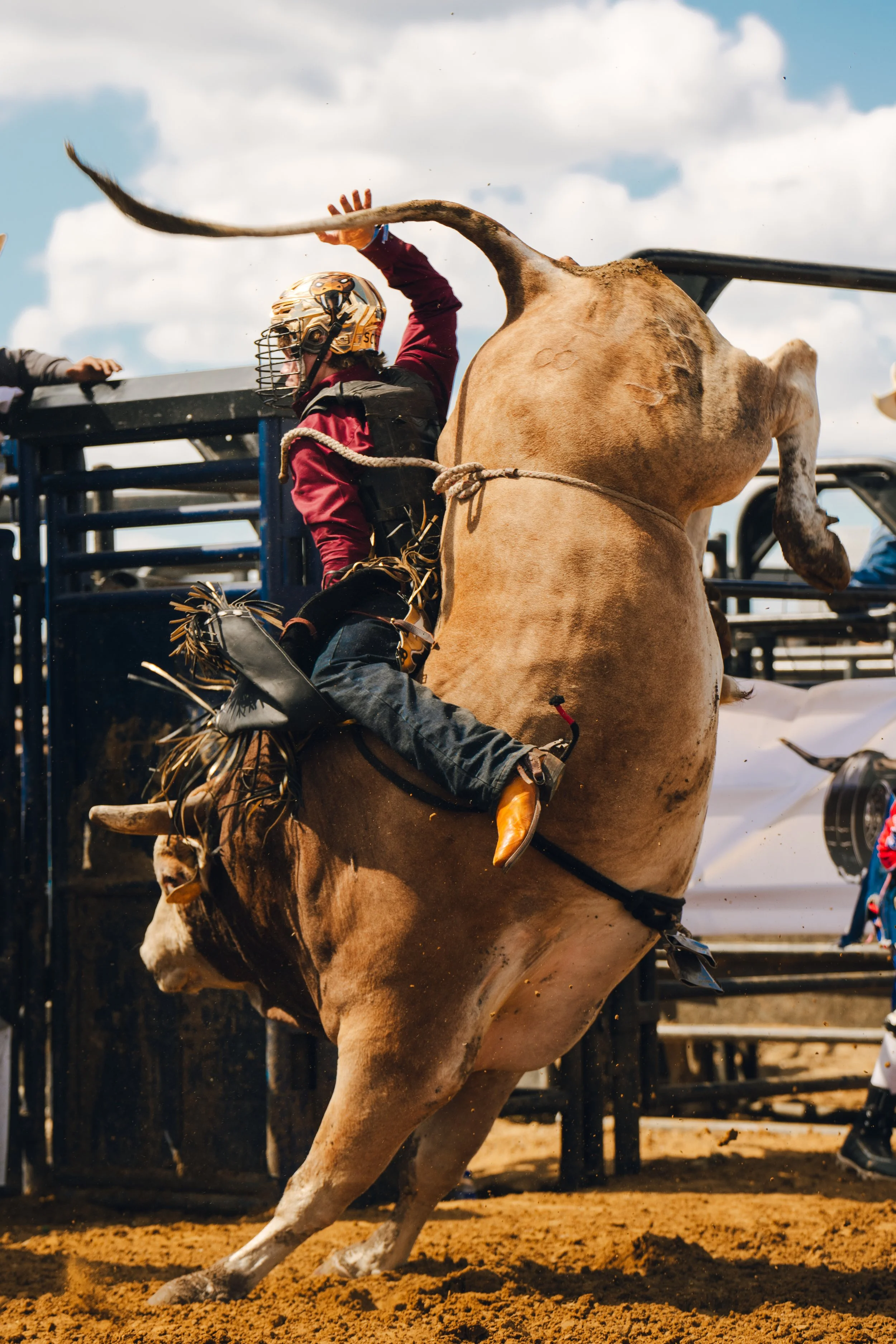 A rodeo rider on a bucking horse with clouds in the background and rodeo equipment visible.