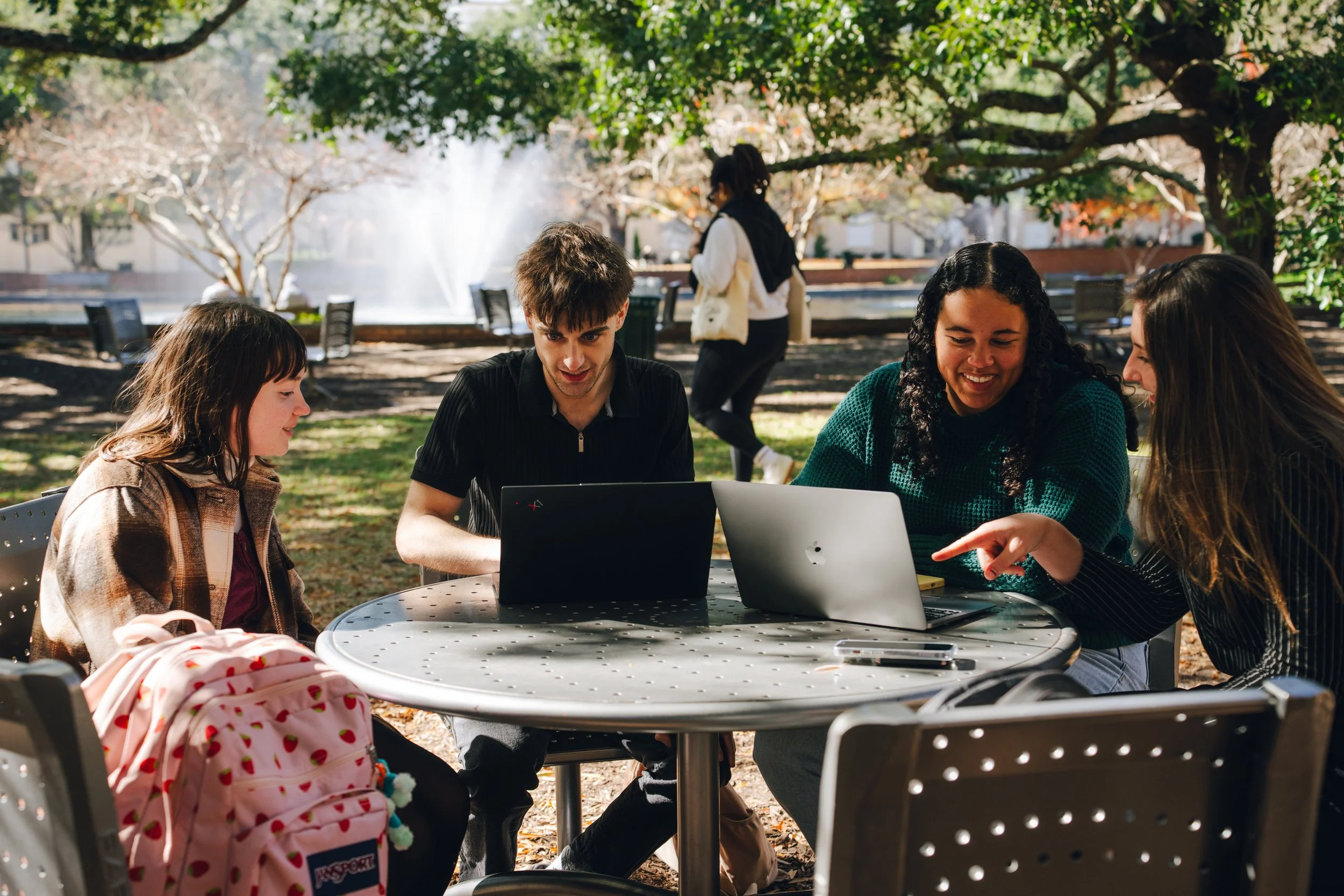 Group of young people sitting around a table outdoors at a park, using laptops and talking, with trees and a fountain in the background.