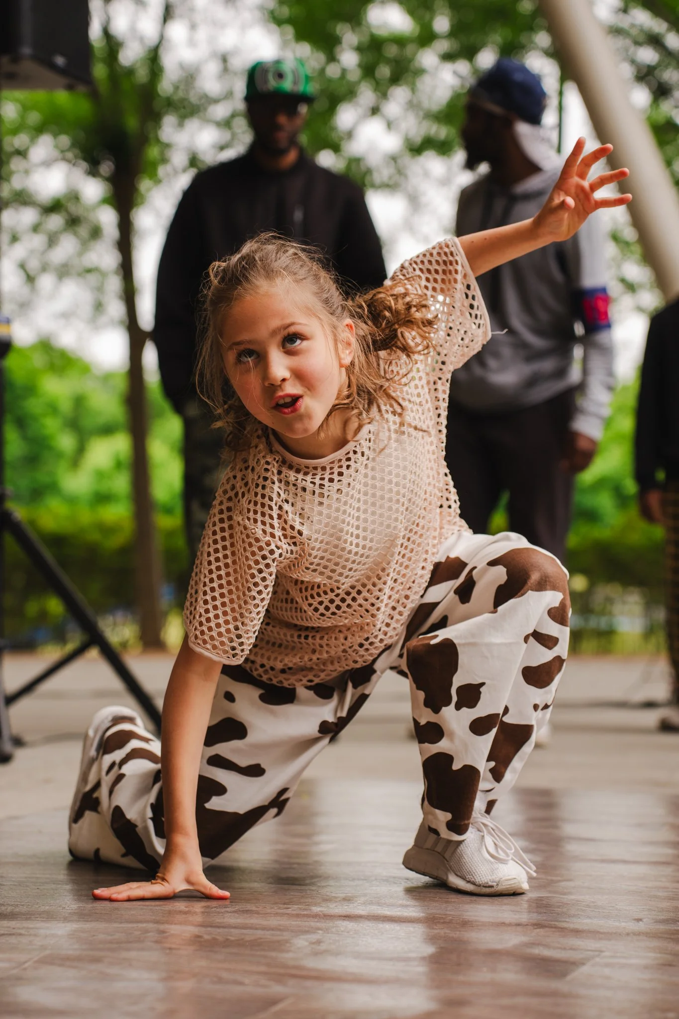 A young girl with curly hair performing a dance move on a wooden floor, wearing a beige mesh top, cow print pants, and white sneakers, with two men standing in the background in an outdoor park setting.