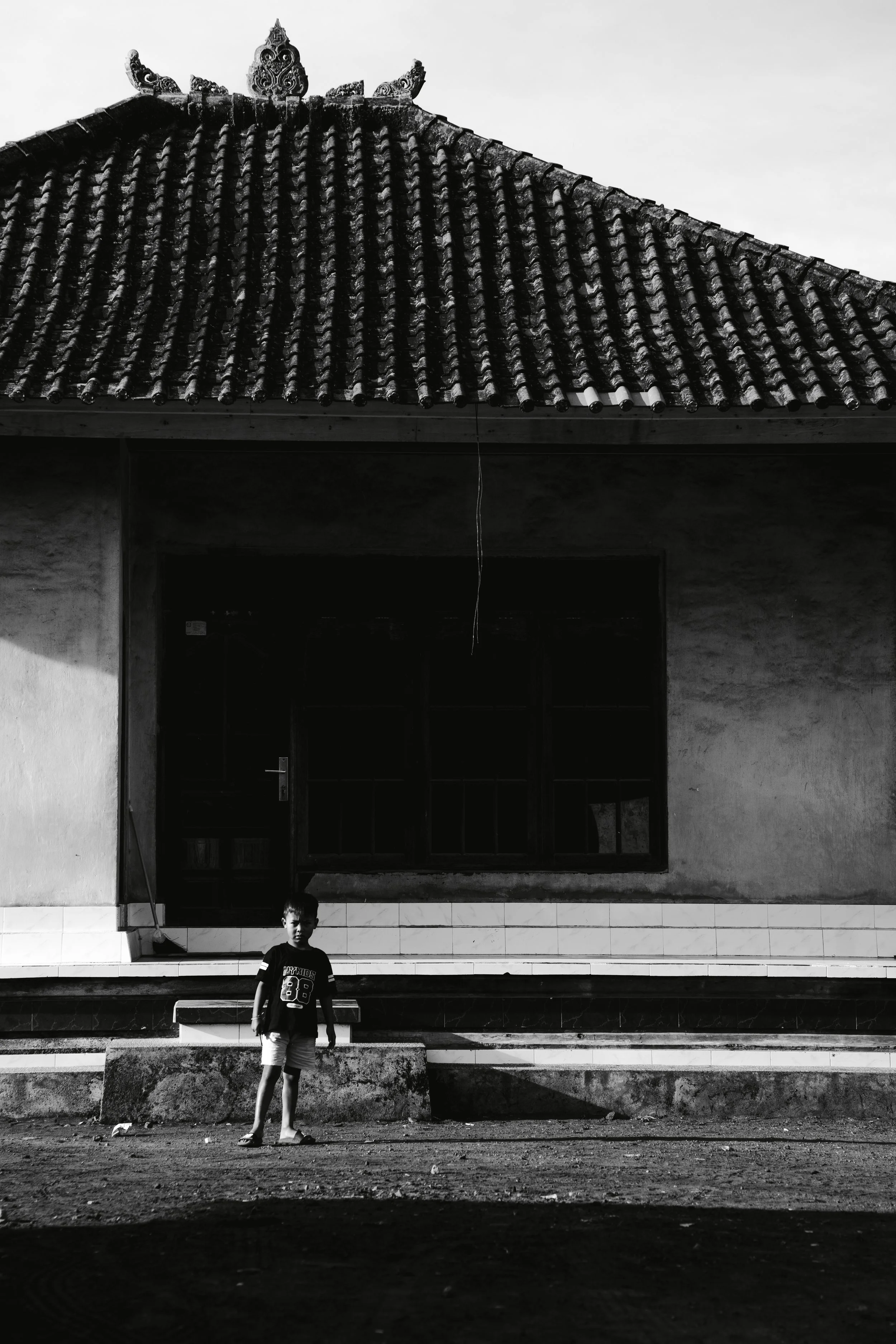 A black-and-white photo of a building with traditional Asian-style roof at the top. In front of the building, a young boy is standing on the stairs, looking at the camera. The ground in front is uneven with some debris.