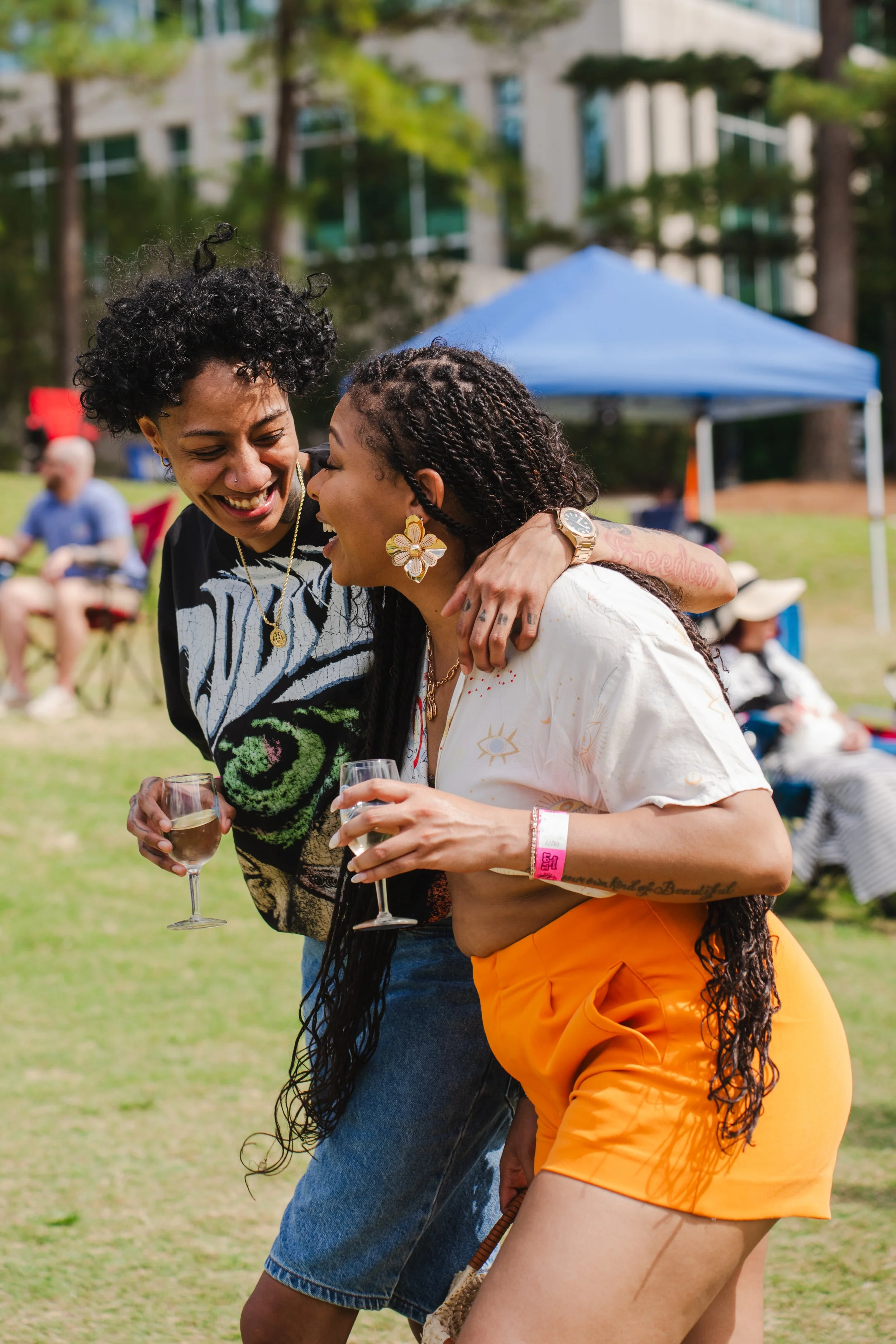 Two women enjoying an outdoor event, laughing and holding glasses of champagne, with a park, other people, and a blue canopy in the background.