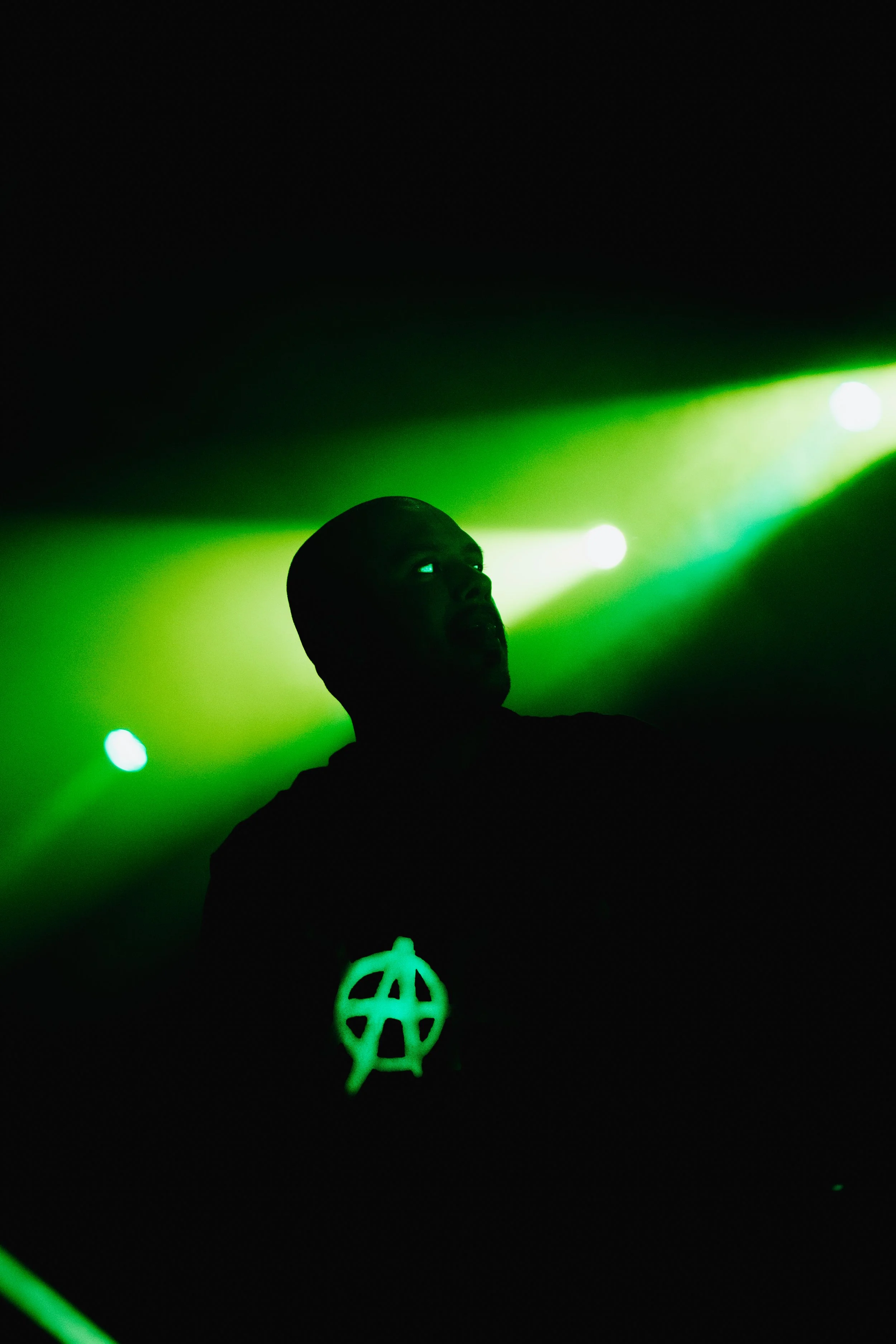 Person with shaved head illuminated by green light, wearing a necklace with peace symbol pendant, standing in a dark environment with bright green lights in the background.