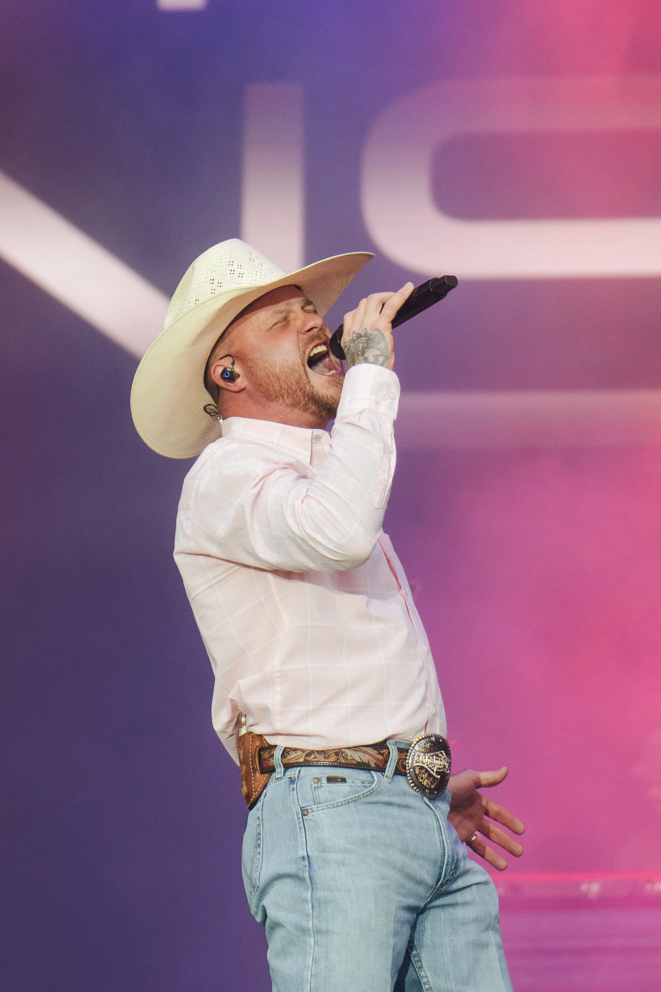 A man in a cowboy hat singing into a microphone on stage, wearing a light plaid shirt and jeans, with a large belt buckle, against a colorful background.