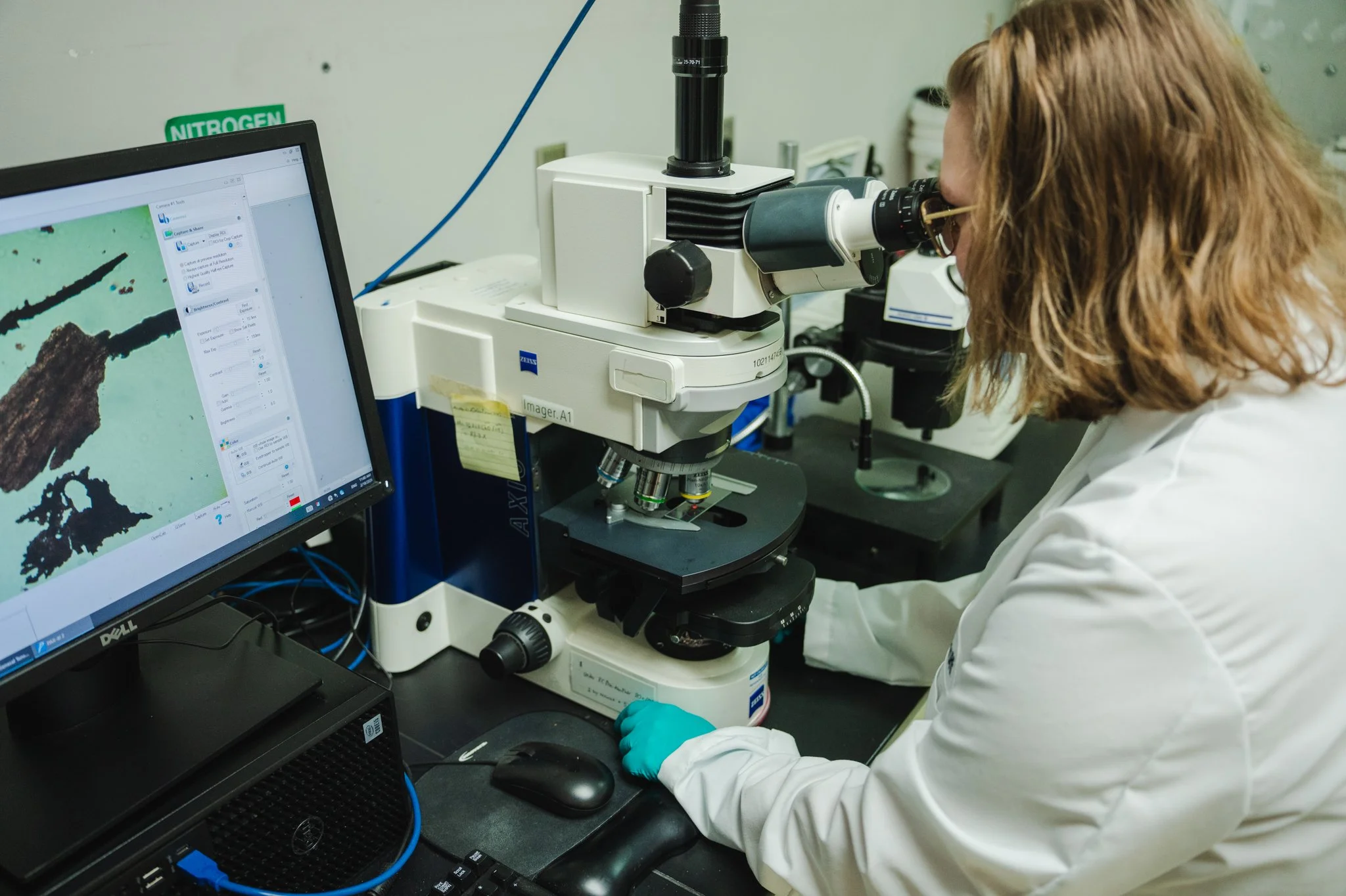 A scientist with red hair, glasses, and blue gloves using a microscope in a lab. A computer monitor displays a magnified image next to the microscope, and a green "NITROGEN" sign is on the wall.