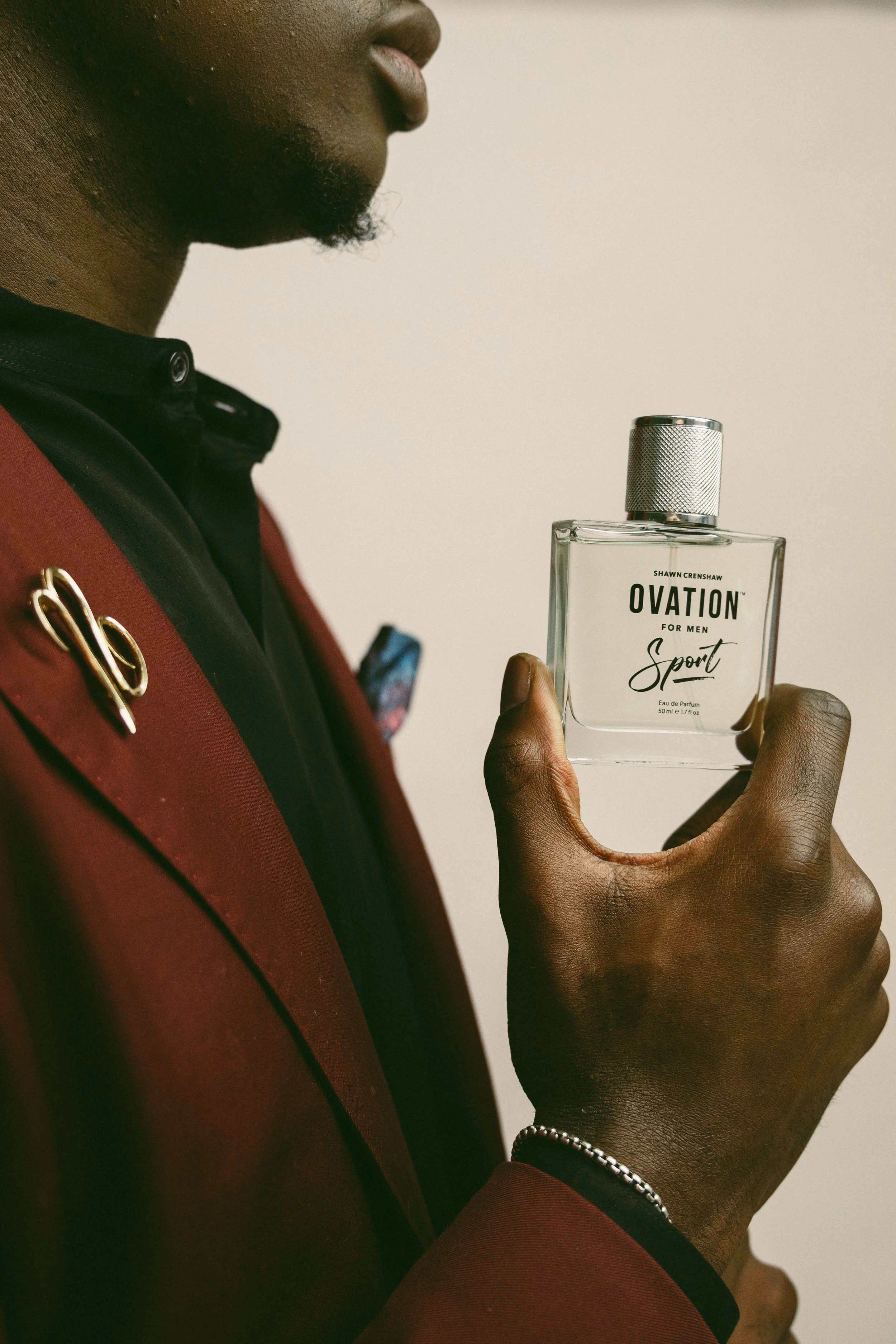 Close-up of a person holding a bottle of Shawn Crenshaw Ovation Sport perfume, with part of their face and hand visible, wearing a red jacket and a silver bracelet.