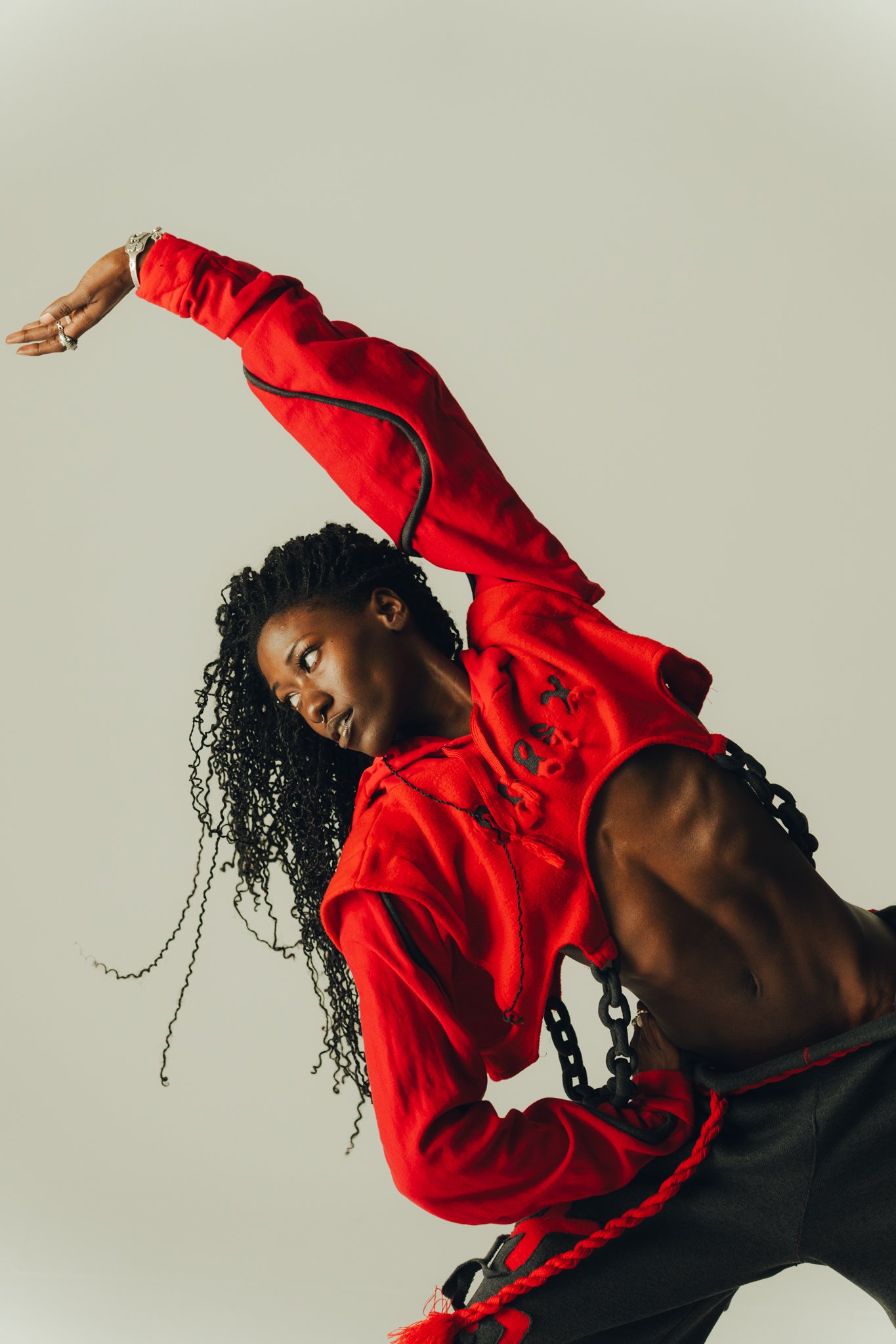 A woman with curly hair dressed in a red jacket and black pants, in a dance pose against a plain light-colored background.