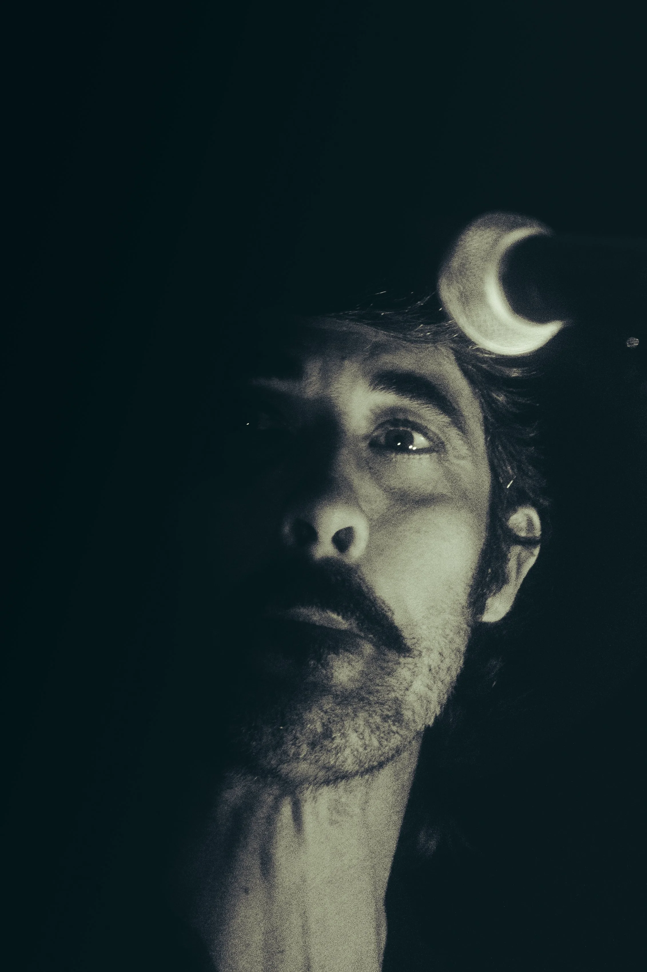 A black and white photograph of a person's face, with only the upper part of their face visible, and the person looking upwards. The person's eye, eyebrow, forehead, part of the nose, and some facial hair can be seen. The background is dark, and a cr