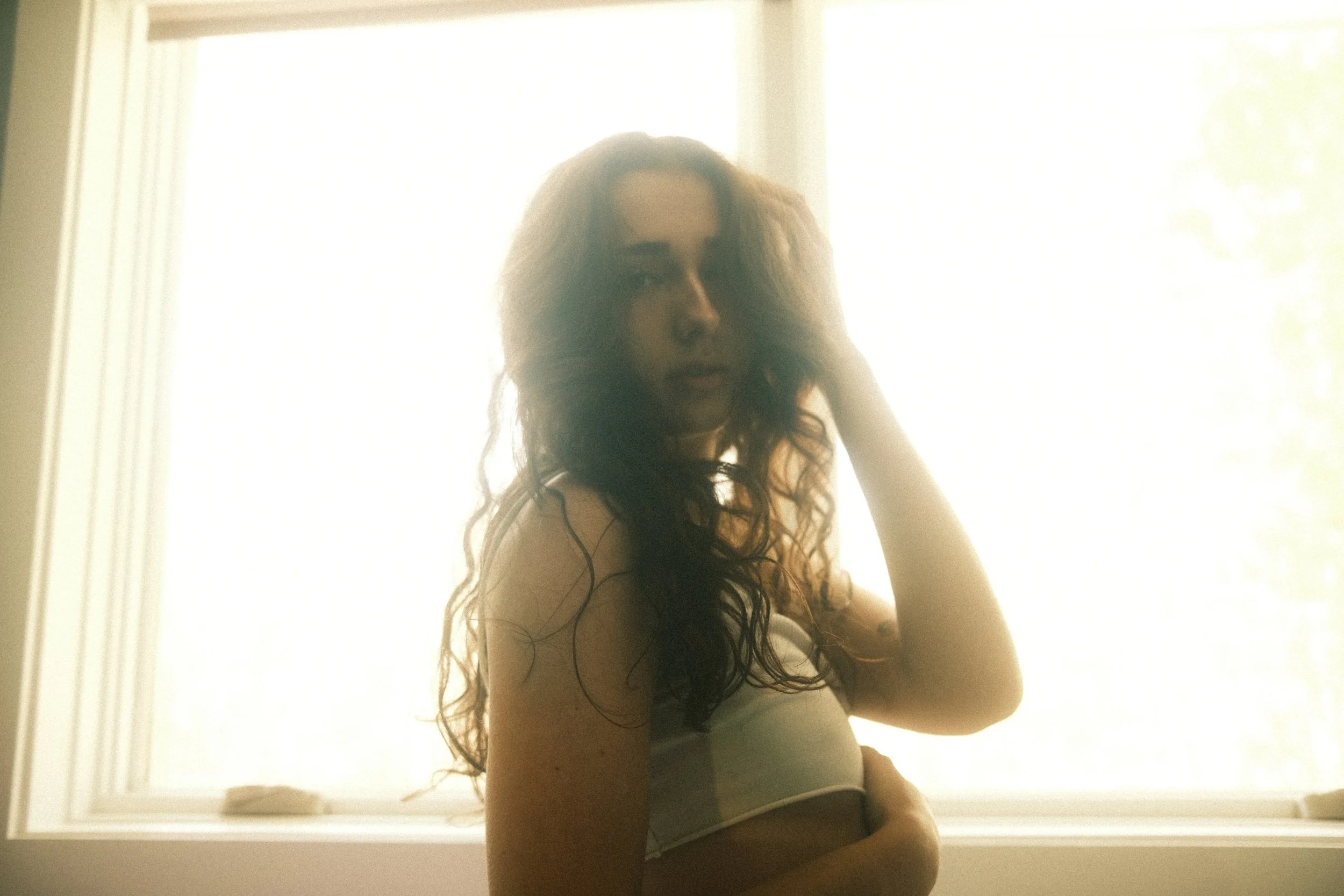 A woman with long curly hair sitting by a window, backlit by sunlight, wearing a tank top and underwear.