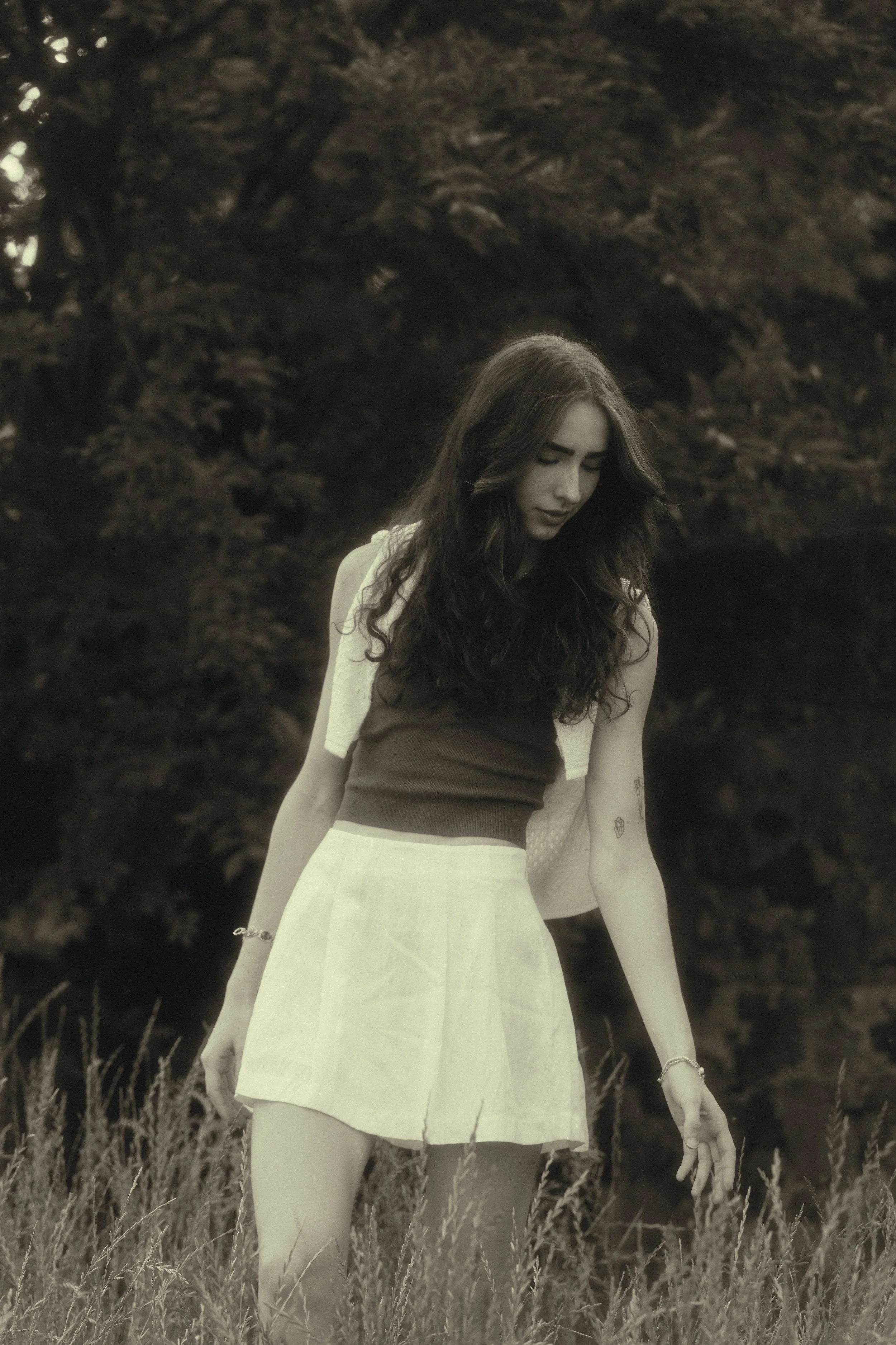 A young woman with long curly hair, wearing a sleeveless dress, standing in a field with tall grass and trees in the background, looking down with a contemplative expression.