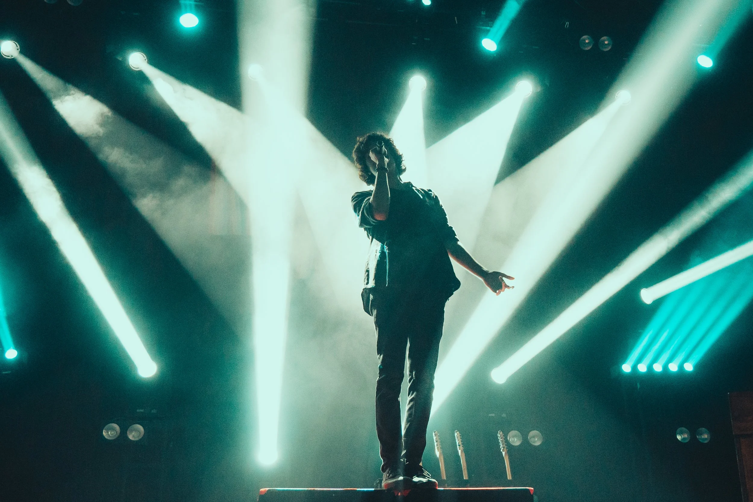 A performer on stage singing into a microphone with bright stage lights shining behind them.