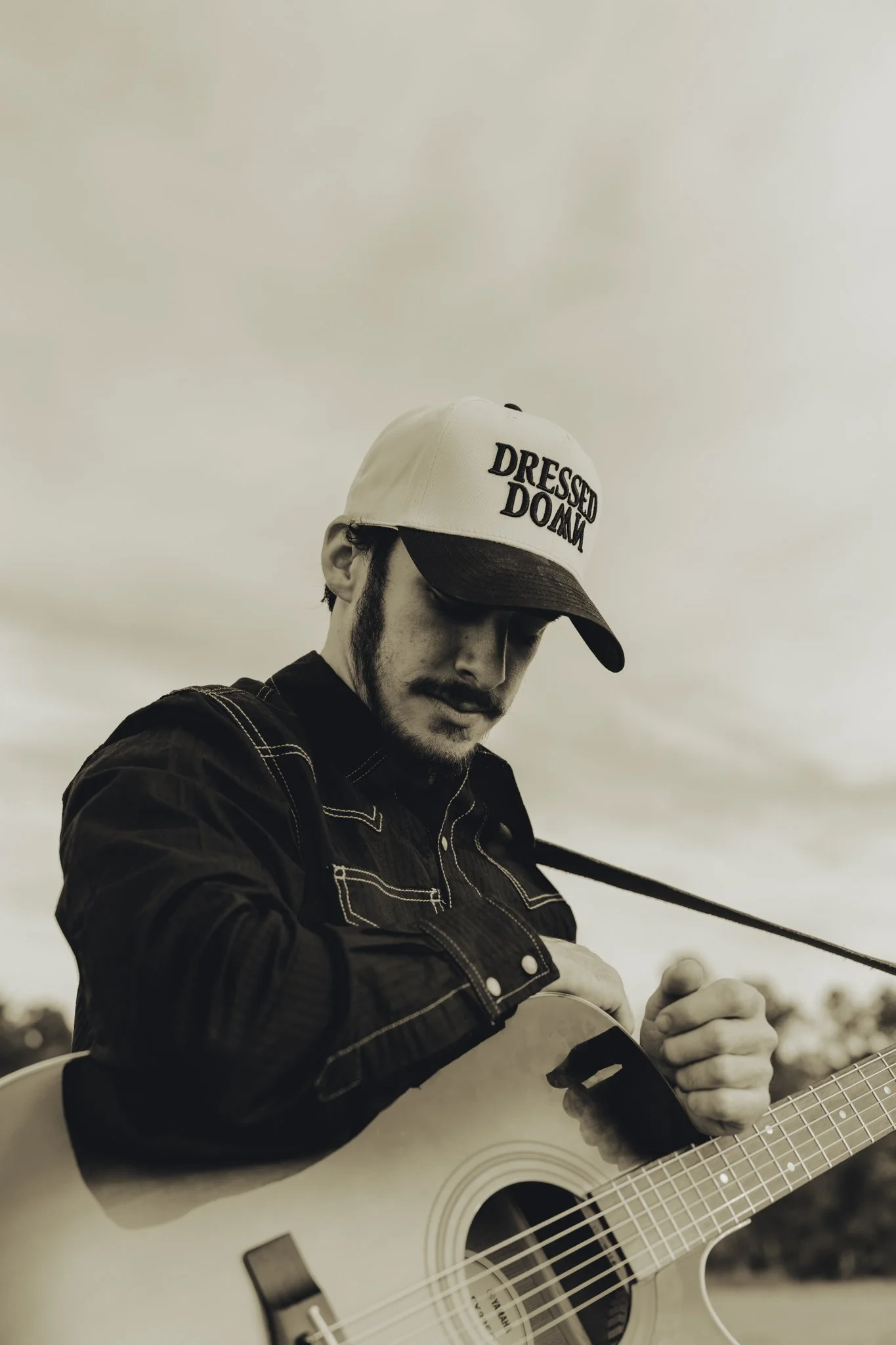 A black-and-white photo of a man playing an acoustic guitar. He is wearing a baseball cap with the words 'DRESSED DOWN' and a casual jacket. The man's face is partially visible, with a focus on his relaxed posture and the guitar.