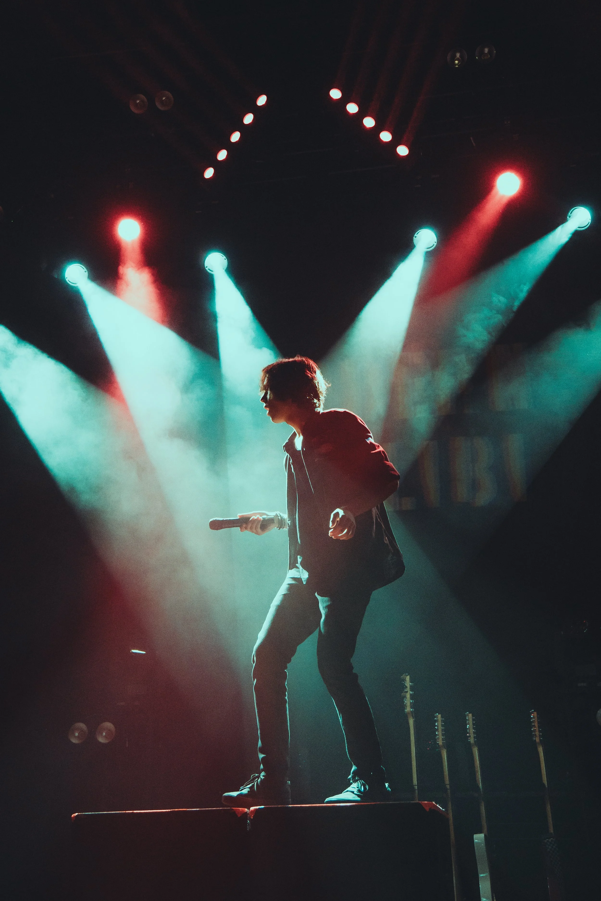 A person on stage holding a microphone, standing on a small platform with colorful stage lights overhead creating a dramatic effect.
